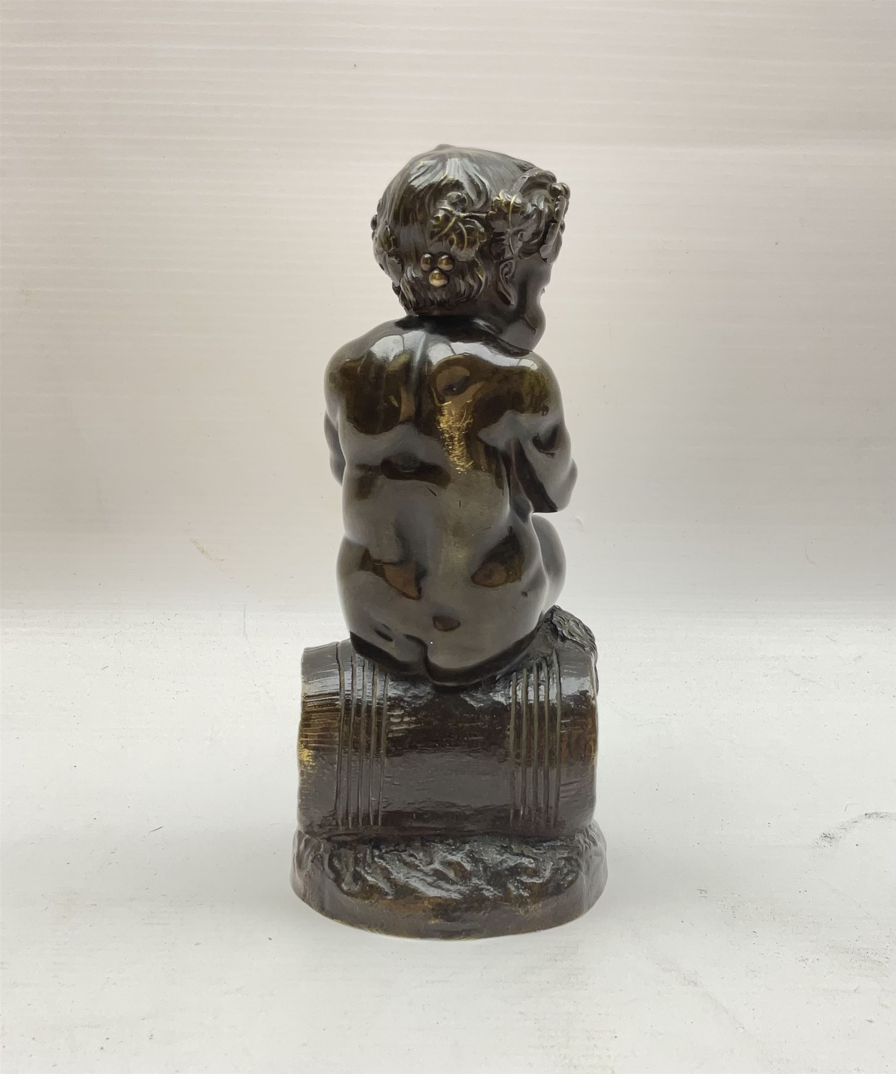 Bronzed metal model of a putti seated upon a barrel with goblet in hands, and fruiting vines adorning their head, H17.5cm