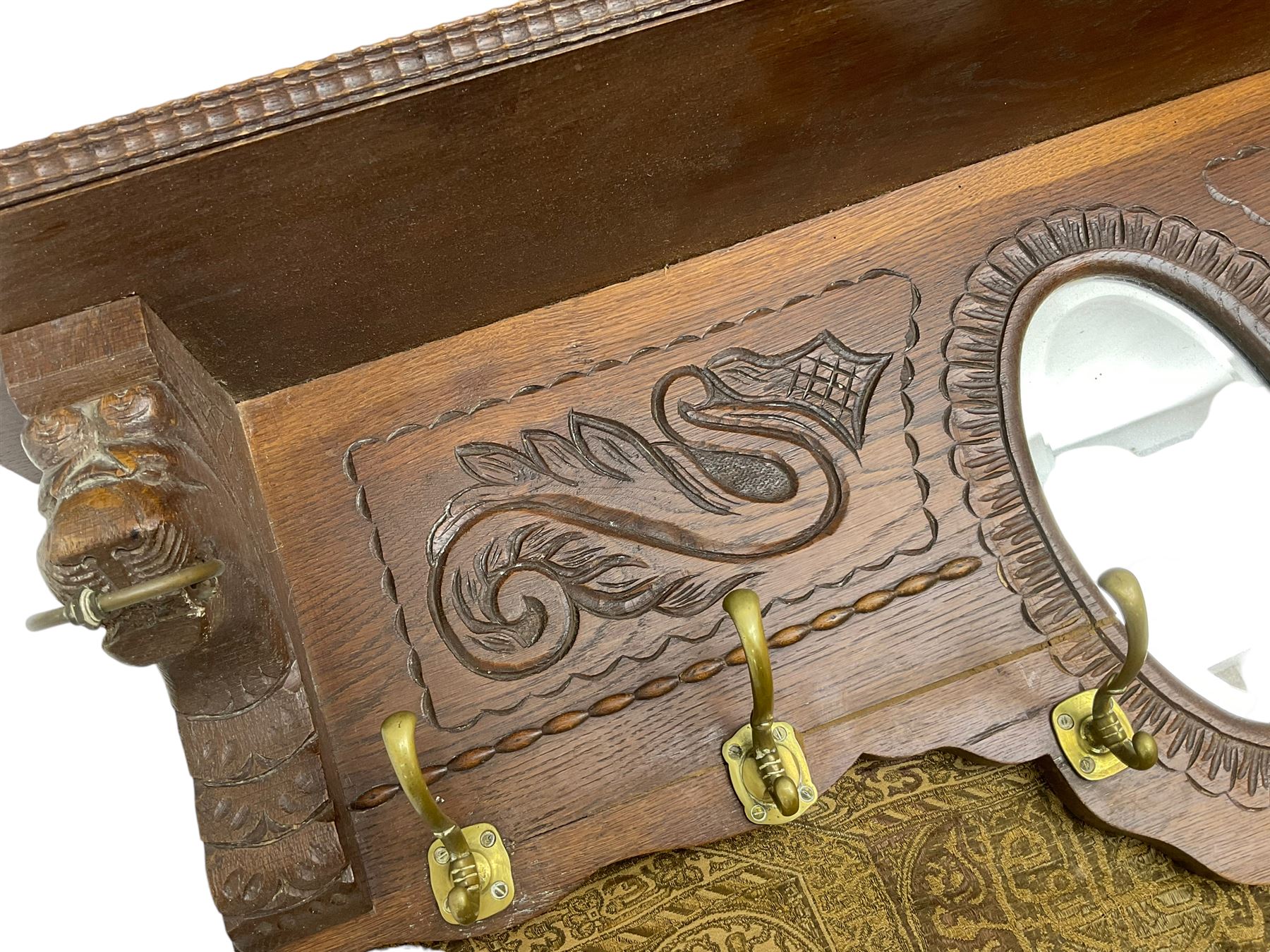 Late 20th century oak wall hanging coat rack, central bevelled oval mirror in foliate carved surround, decorated with S-scrolls and lion masks, fitted with needlework hanging 