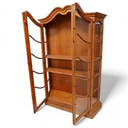 Polished pine display cabinet, shaped top over two glazed doors, fitted with three shelves, on moulded plinth base 