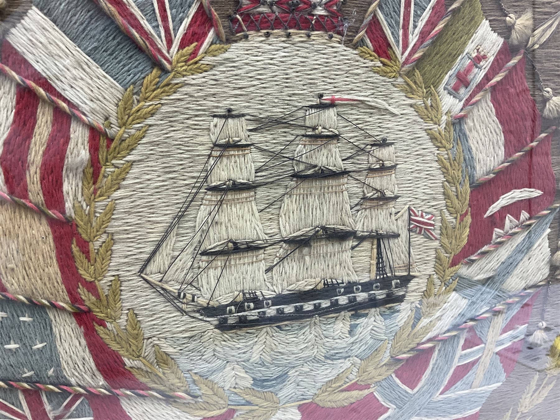 19th century woolwork, depicting a clipper ship, fully rigged over black hull with white ports, within a border of ensigns and flags and beaded crown above, in glazed wooden frame, H57cm