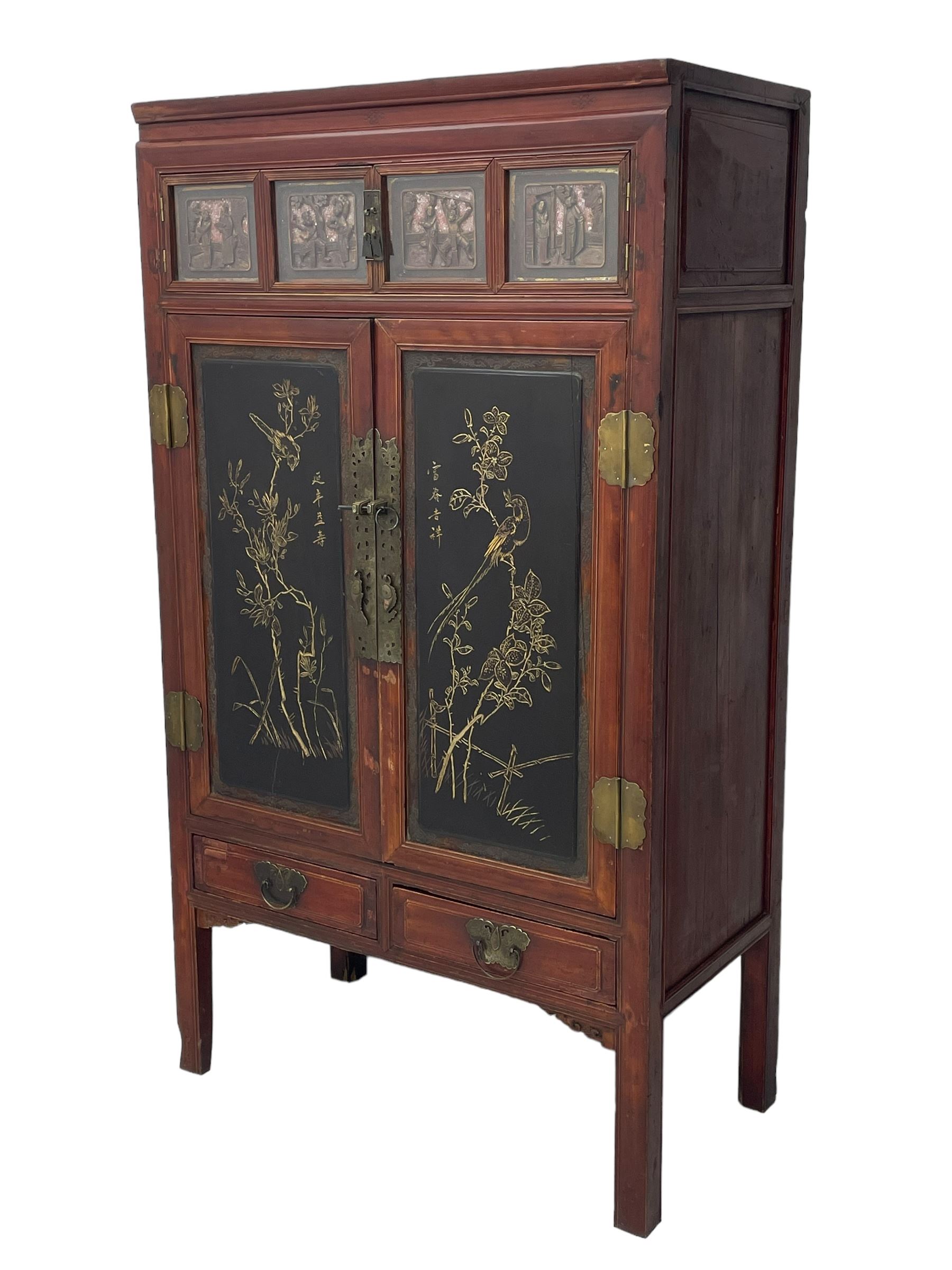 Late 19th century Chinese Qing dynasty red and black lacquered wood cabinet, Fujianese province, upper cupboard enclosed by two doors with relief carved and gilt panels depicting figures behind glass, the large cupboard enclosed by two panelled doors carved with trailing foliate branches and birds, inscribed with Chinese characters, to the left ‘Prolong life’ and to the right ‘Wealth and good fortune’, fitted with two drawers, square supports with carved brackets 