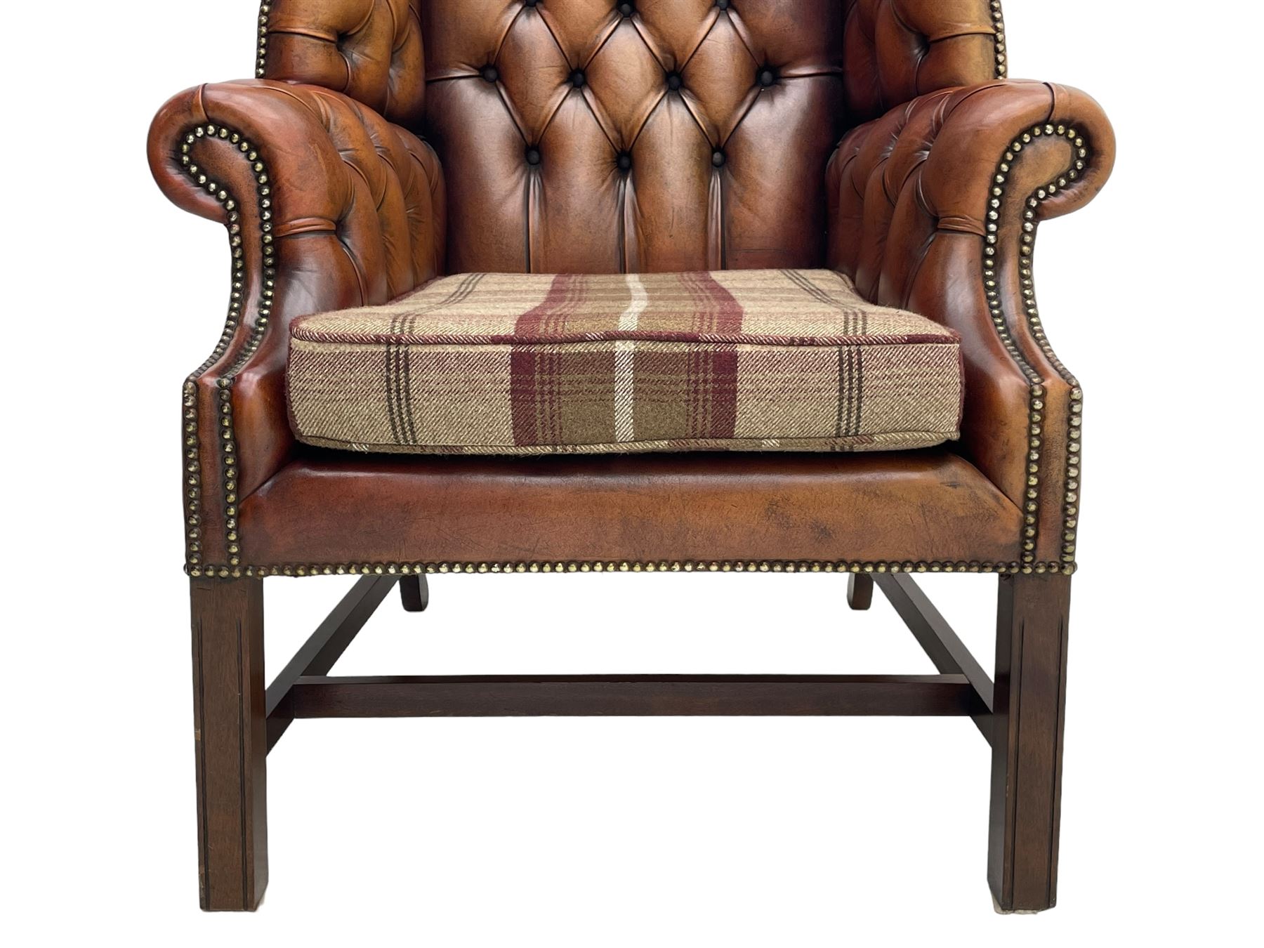 Georgian design hardwood-framed wingback armchair, shaped cresting rail and deep wingback over rolled arms, upholstered in brown buttoned leather with studwork bands, the seat cushion upholstered in checkered fabric, on square supports united by stretchers (W86cm, H118cm, D86cm); together with matching rectangular footstool (63cm x 43cm, H37cm) 