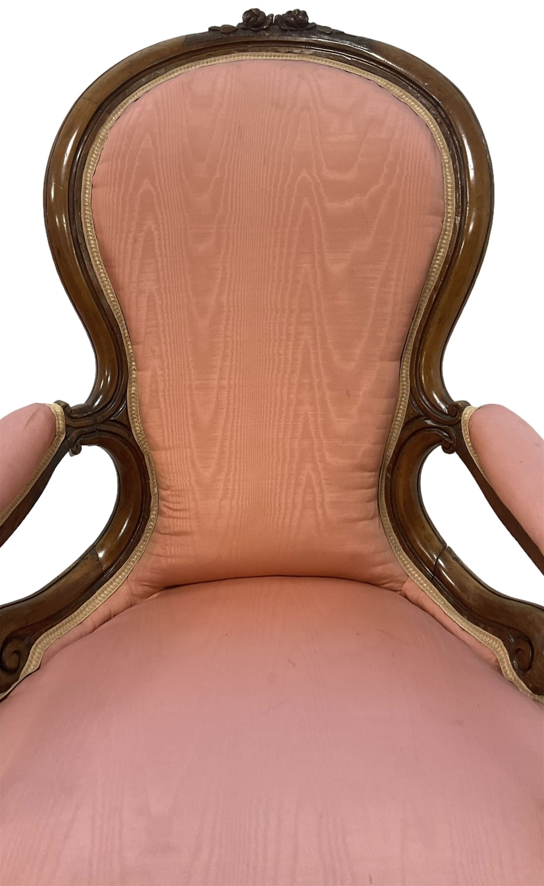 Victorian mahogany framed open armchair, spoon-back and sprung seat upholstered in pink fabric, scrolled arm terminals over serpentine fronted seat decorated with moulded carved floral motifs, cabriole supports with ceramic castors