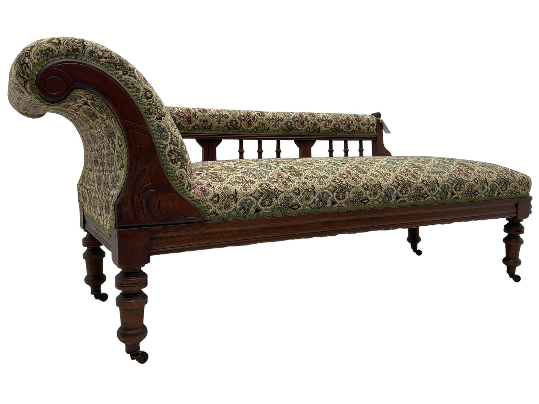 Late 19th century walnut framed chaise longue, upholstered in floral pattern fabric, rolled back rest and turned balustrade back, on turned feet with brass and ceramic castors 