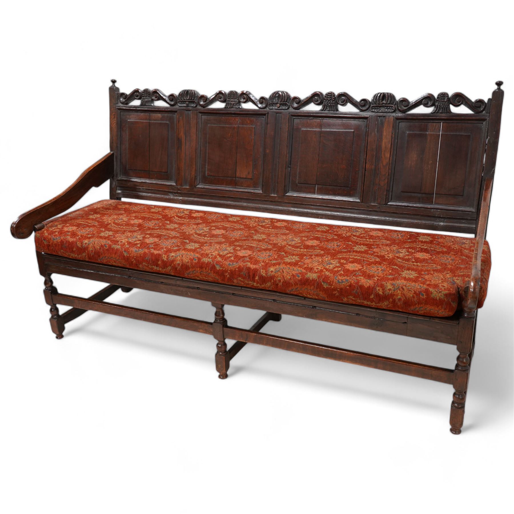 Early 18th century oak hall bench or settle, the cresting rail carved with S-scrolls and crown motifs, over a quadruple panelled back with carved rail, loose seat cushion upholstered in terracotta foliate fabric, flanked by shaped arm terminals, raised on turned supports united by stretchers