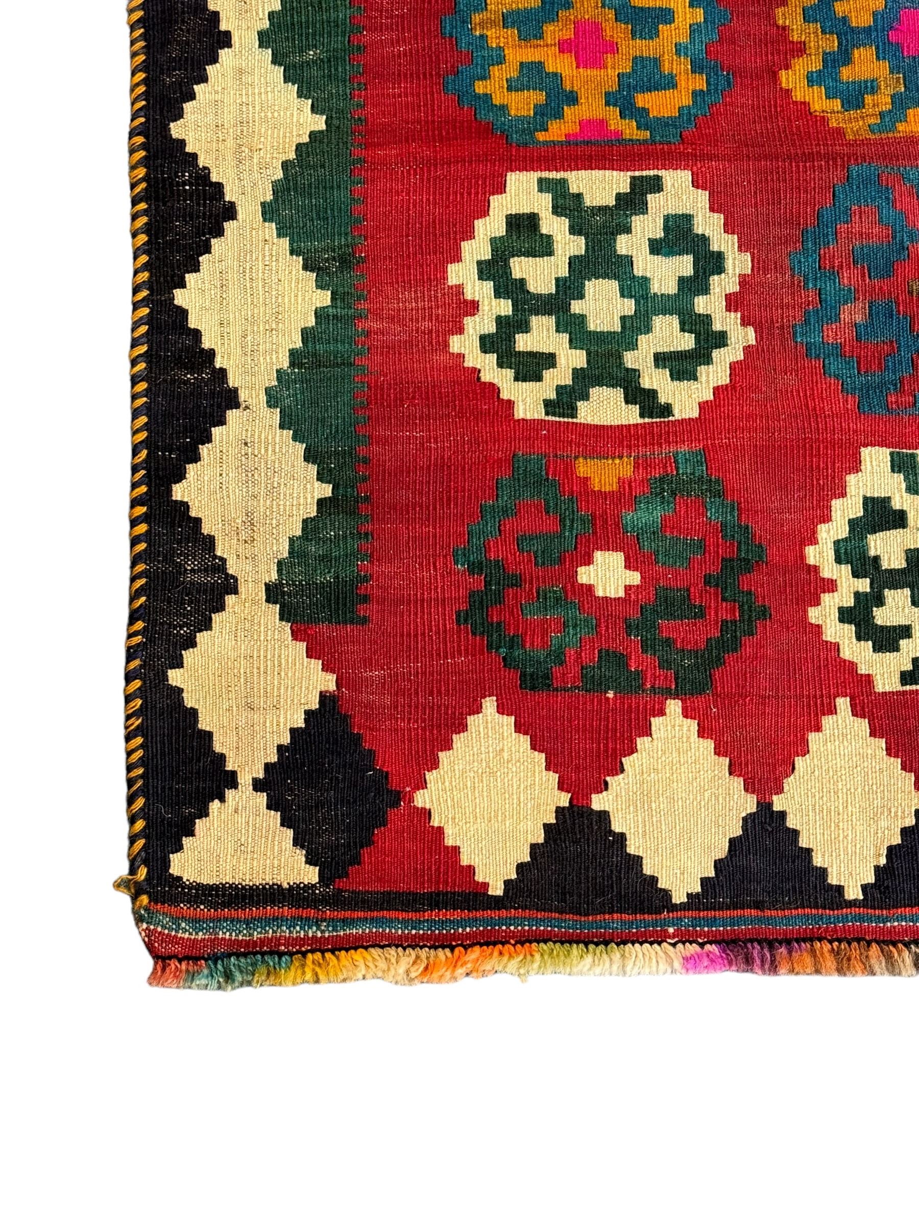 Southwest Persian Qashqai Kilim red ground rug, the central field adorned with rows of multicolored hexagonal medallions with geometric patterns, the border featuring alternating black and white stepped diamonds, narrow band to one end decorated with multicoloured geometric figures