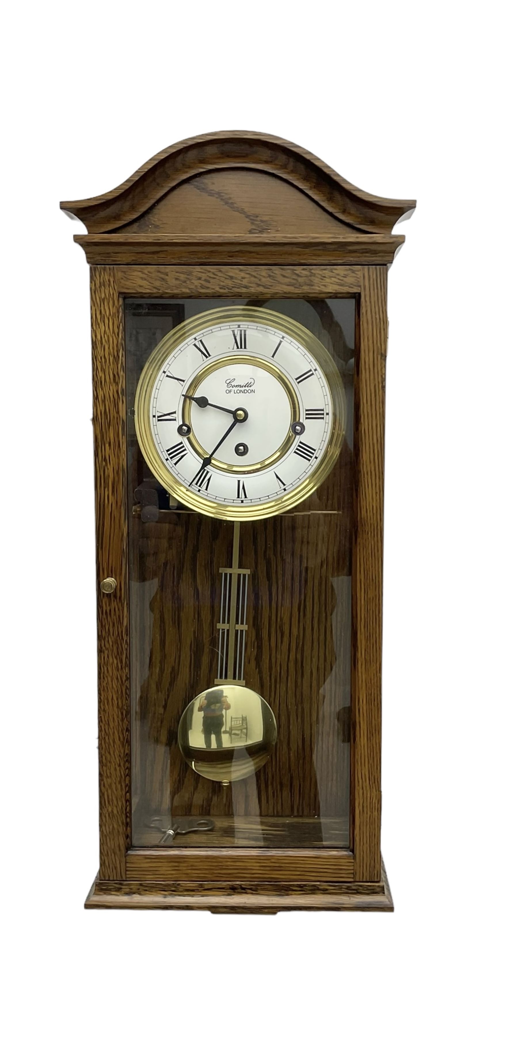 Commiti of London -  three train spring driven 8-day wall clock in an oak case, with a fully glazed door and curved pediment, two part dial with Roman numerals, spade hands and gridiron pendulum, rack movement chiming the hours and quarters on 5 gong rods. With key.