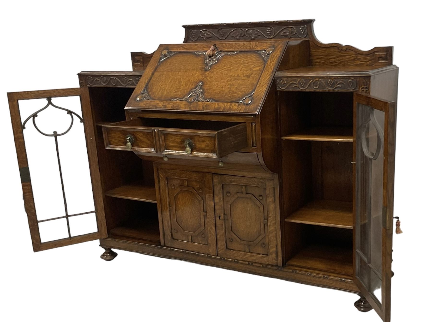 Early 20th century heavily carved oak bookcase bureau, raised back with grape-vine carved frieze, over fall-front enclosing fitted interior, above two short drawers and curved frieze drawer, flanked by astragal glazed and panelled cupboards, on turned feet