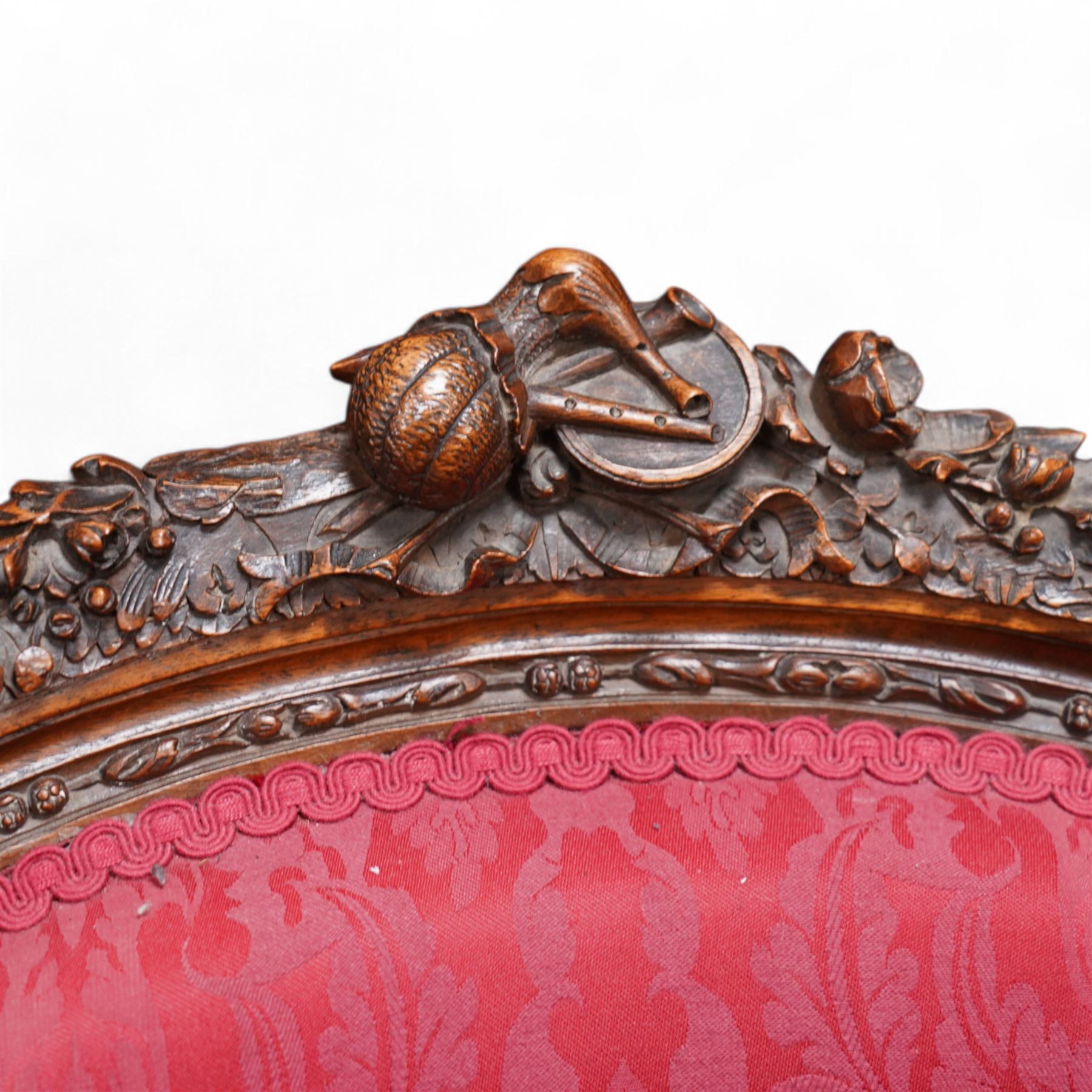 Late 19th century walnut framed fauteuil armchair, arched cresting rail carved with musical instruments and extending floral decoration, curved wing back with moulded and bead carved frame, upholstered in crimson fabric with repeating foliate pattern, acanthus carved arm terminals over acanthus and ribbon carved arm supports, the seat rail carved with fruiting foliage band, on turned and stop-fluted feet 