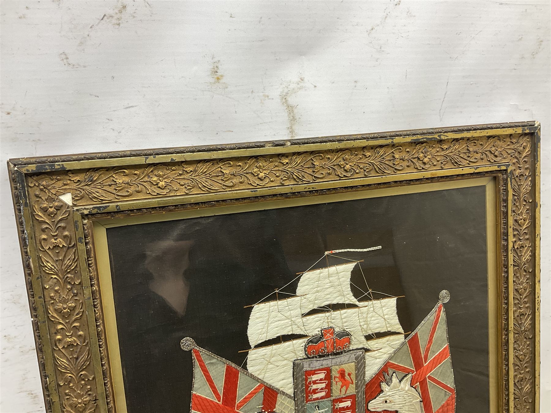 Early 20th century silk needlework panel, depicting a ship in full sail and Royal Coat of Arms to centre, with red and white ensigns to either side, lion and unicorn below with quote 'Dieu Et Mon Droit', upon a black silk ground within gilt frame, H59cm