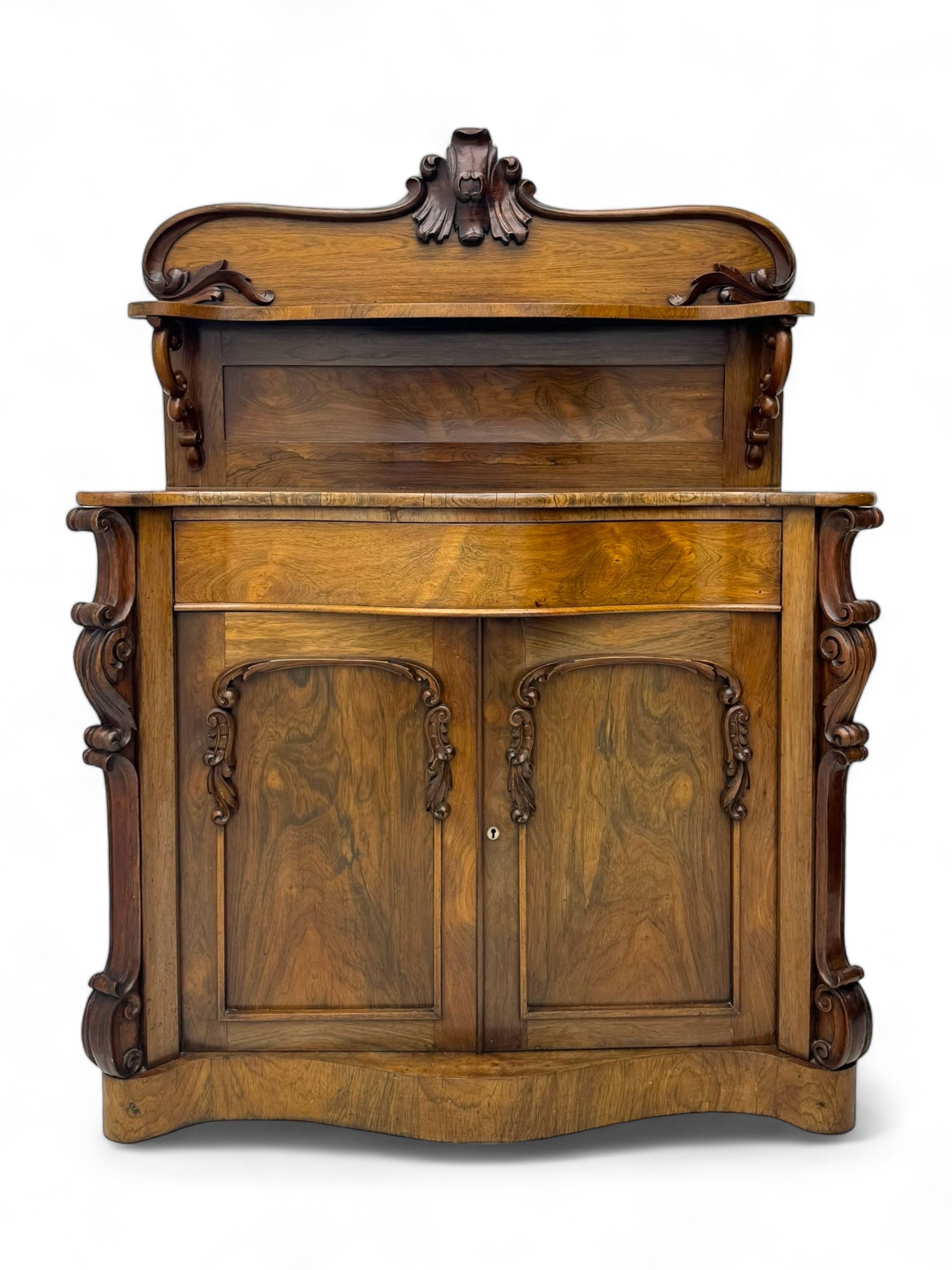 Mid-19th century rosewood chiffonier, raised back with scroll and leaf carved shaped pediment, serpentine top over drawer and double cupboard with scrollwork decoration, on shaped plinth base