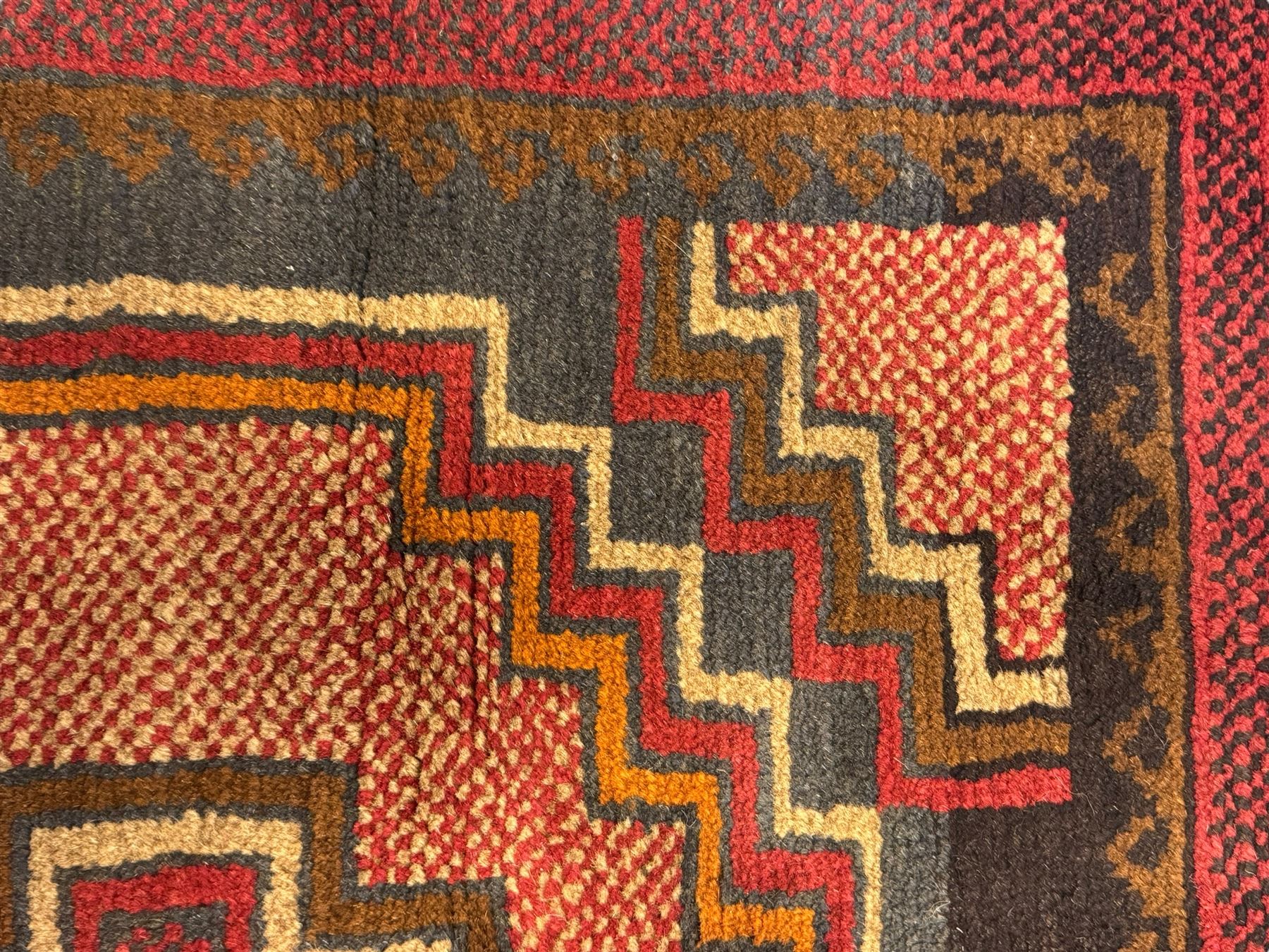 Baluchi crimson ground rug, the central field decorated with two stacked geometric lozenge medallions, surrounded by stepped multicoloured motifs, the wide border with alternating angular motifs in deep red, within a narrow brown guard stripe