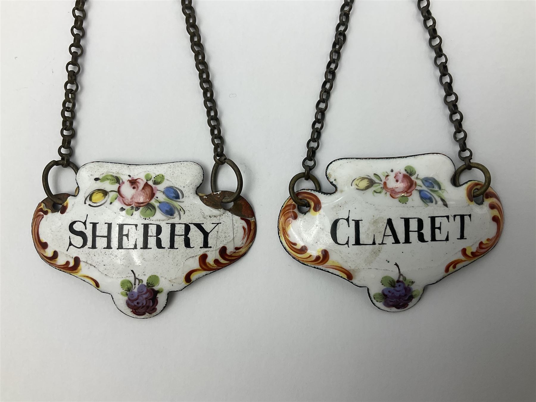 Two late 18th/early 19th century enamel wine labels, each of shaped form, titled 'CLARET', and 'SHERRY' and decorated with floral sprigs and scroll detail upon a white ground, each with suspension chain, each approximately H3.5cm W5cm