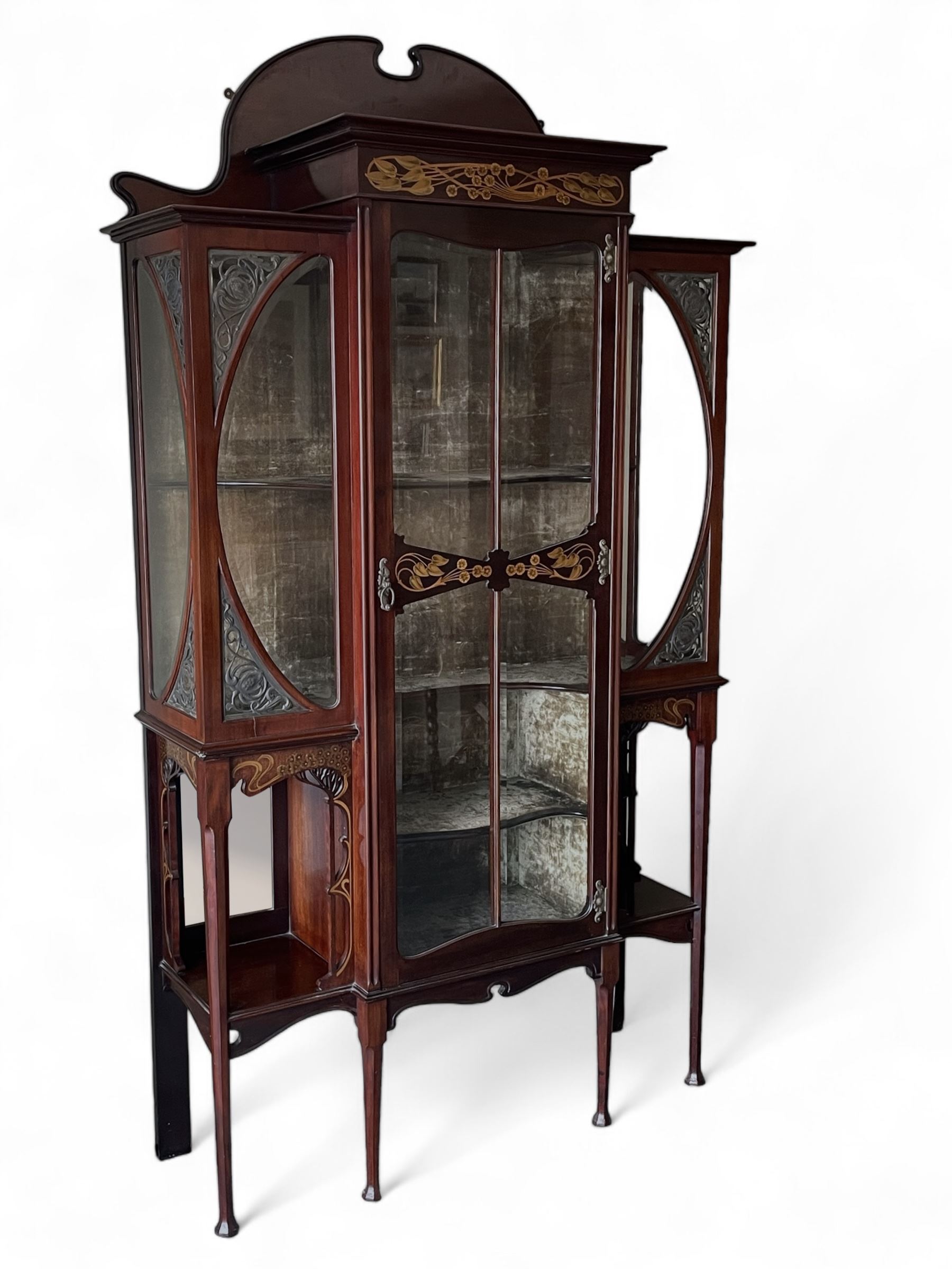 Art Nouveau period inlaid mahogany display cabinet, raised shaped and moulded pediment over projecting moulded cornices, the upper frieze inlaid with trailing branches and tulips, single astragal and bevel glazed door enclosed velvet lined interior fitted with shelves, flanked by curved glass panes decorated with pierced metal foliate spandrels, lower flanking shelves with mirror backs enclosed within shaped and pierced rails inlaid with flower heads, on tapering octagonal supports terminating to compressed pad feet, the lock stamped 'Salmon Bros, London' 