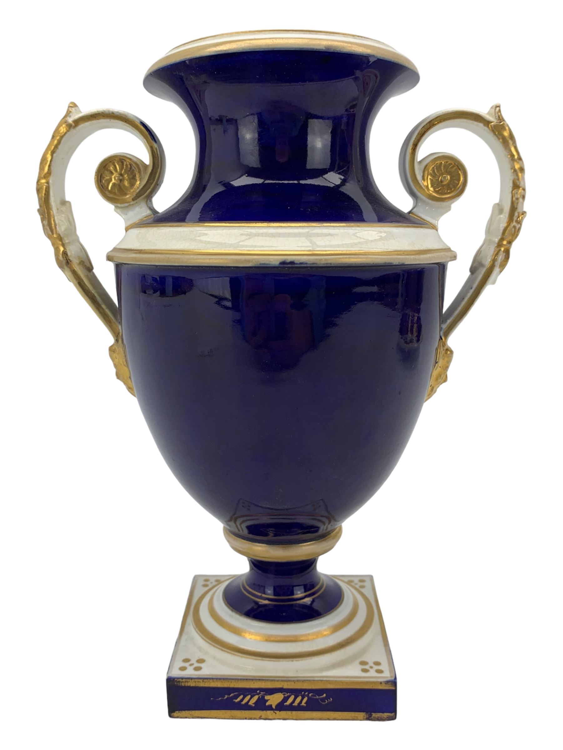 Early 19th century Derby porcelain urn, the reserve painted with a pastoral landscape and castle in the distance, with twin scroll and mask handles, upon a square pedestal base, H23cm, together with a Derby shell-shaped dish, centrally hand painted with a landscape scene titled 'In North Wales', within a cobalt blue and gilt border, L25cm (2)