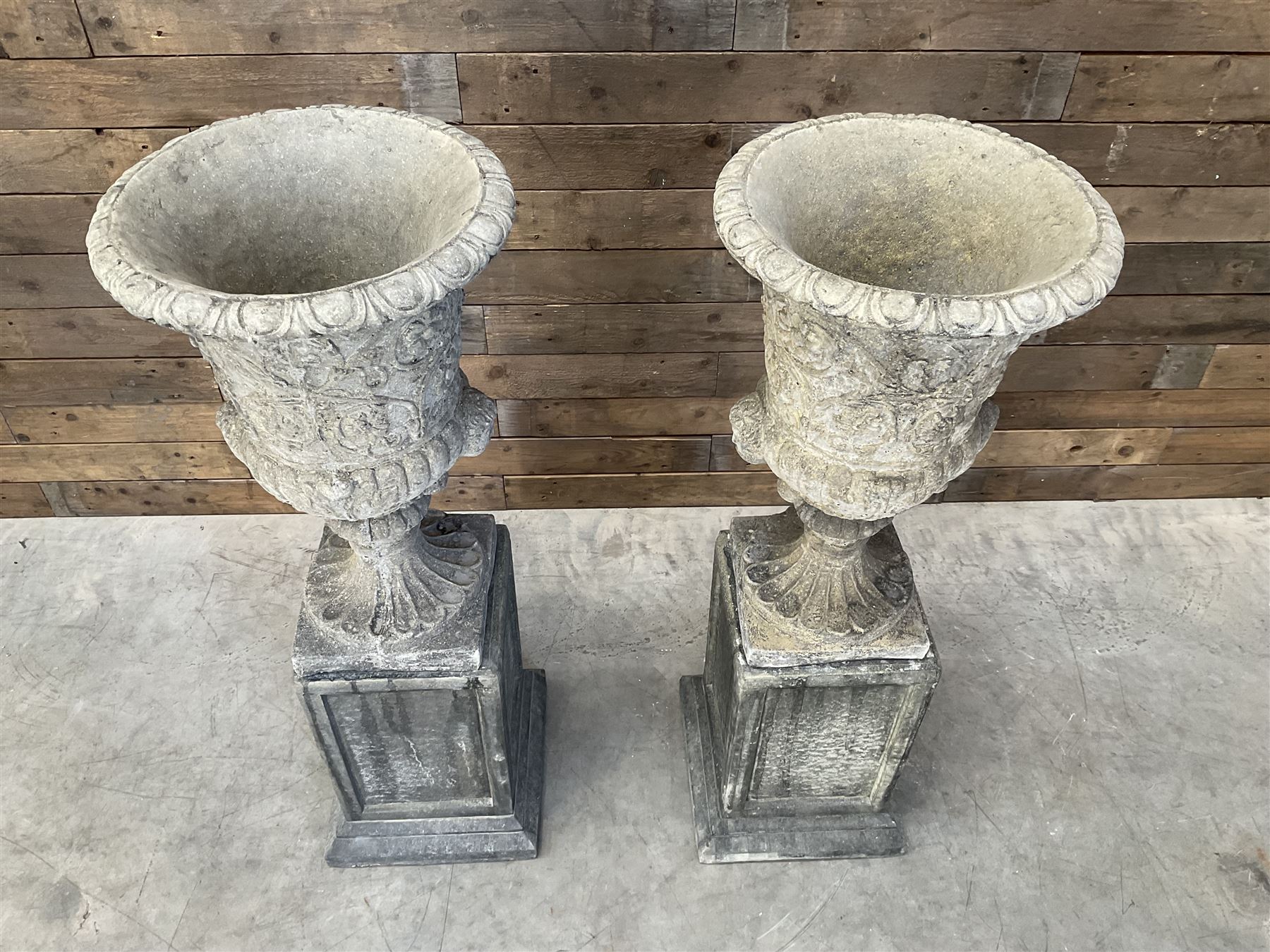 Pair of Georgian design cast stone garden urns, egg and dart rim with raised floral motifs, pedestal base terminating on square plinth, raised on square block plinth