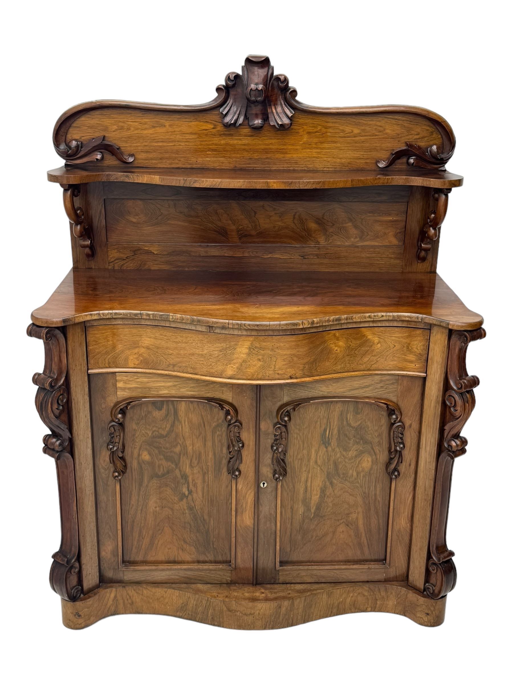 Mid-19th century rosewood chiffonier, raised back with scroll and leaf carved shaped pediment, serpentine top over drawer and double cupboard with scrollwork decoration, on shaped plinth base