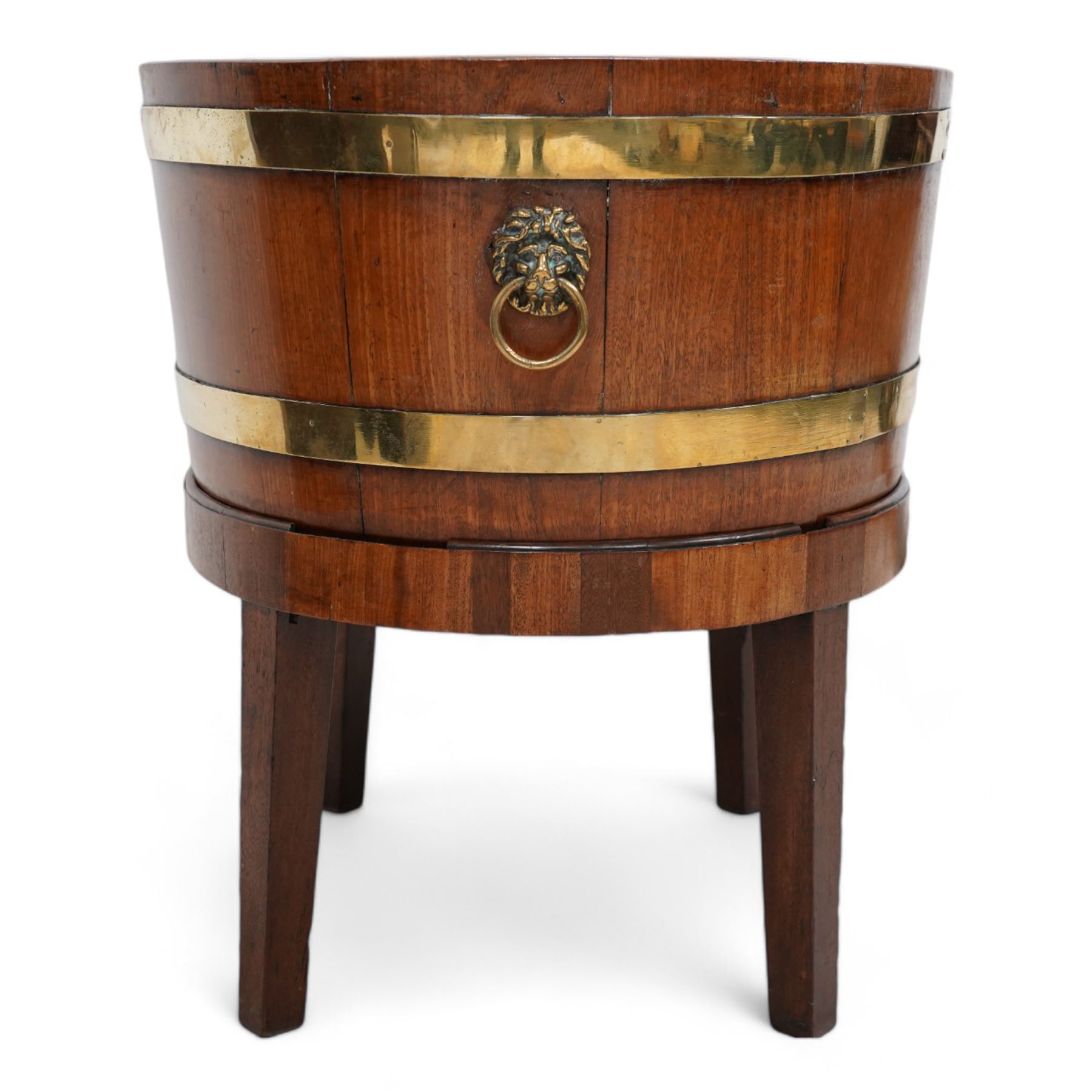 George III Chippendale design brass bound mahogany wine cooler of oval form, the coopered body enclosed by two brass brands, with lead liner to interior, fitted with twin lion mask ring handles, raised on square tapering supports