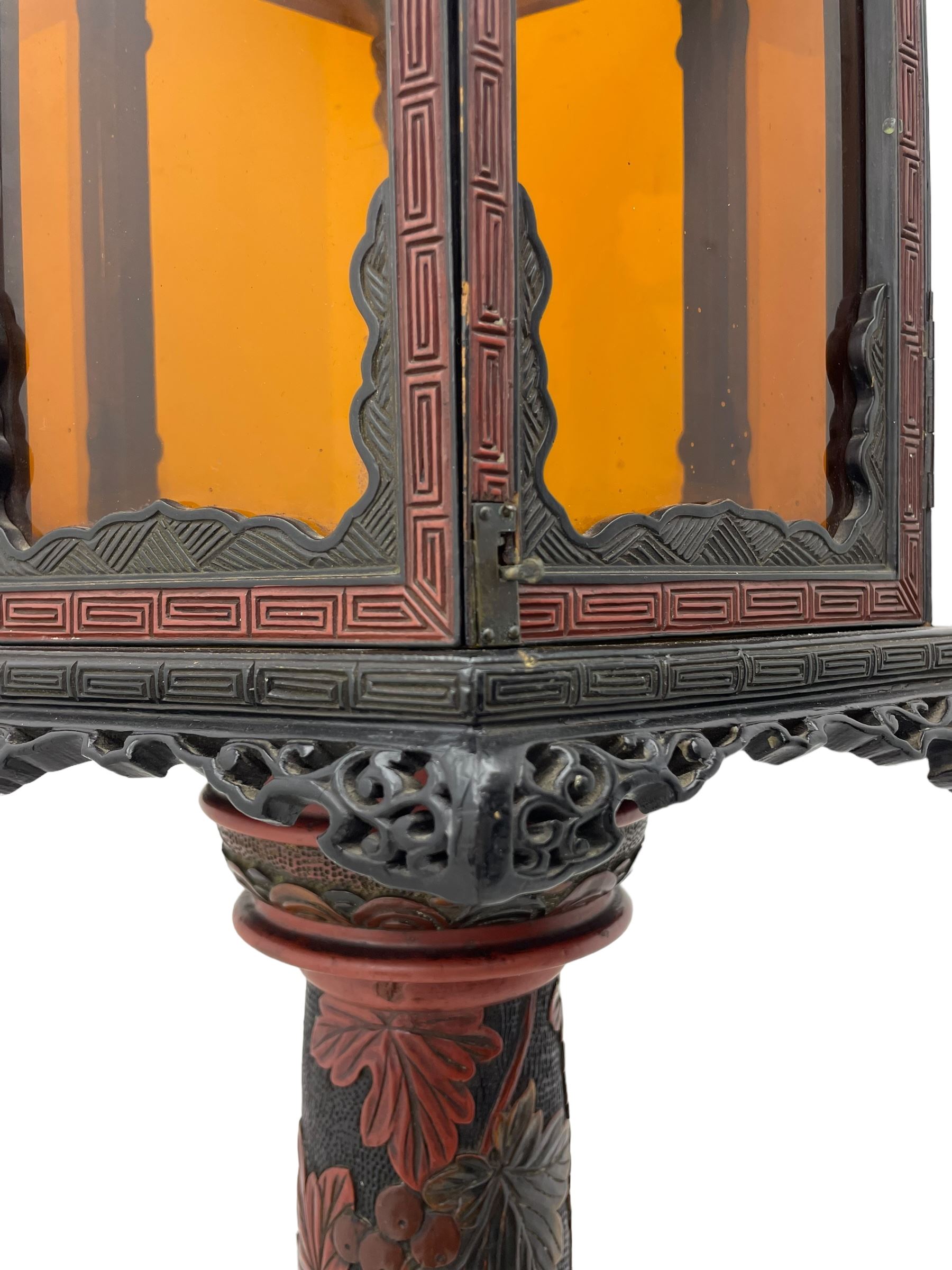 20th century Chinese black and red lacquered hardwood lantern stand, hexagonal shaped canopy top carved with flower heads and scrolls, the lantern fitted with amber tinted glass panes, pierced and carved upper panels, enclosed by two hinged doors, turned stem carved in relief with trailing vine leaves with fruits, stepped hexagonal base with carved decoration  