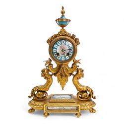 French-mid 19th century 8-day gilt and porcelain mantle clock with a break front plinth raised on paw feet, with recessed rectangular porcelain panels depicting two lovers and floral decoration, drum cased movement with a porcelain dial supported on two matching scroll supports, surmounted with a tapered porcelain urn and gilt finial, dial with cartouche Roman numerals on a blue ground and a depiction of cupid to the dial centre, twin train rack striking movement, striking the hours and half hours on a bell. With Pendulum and key.