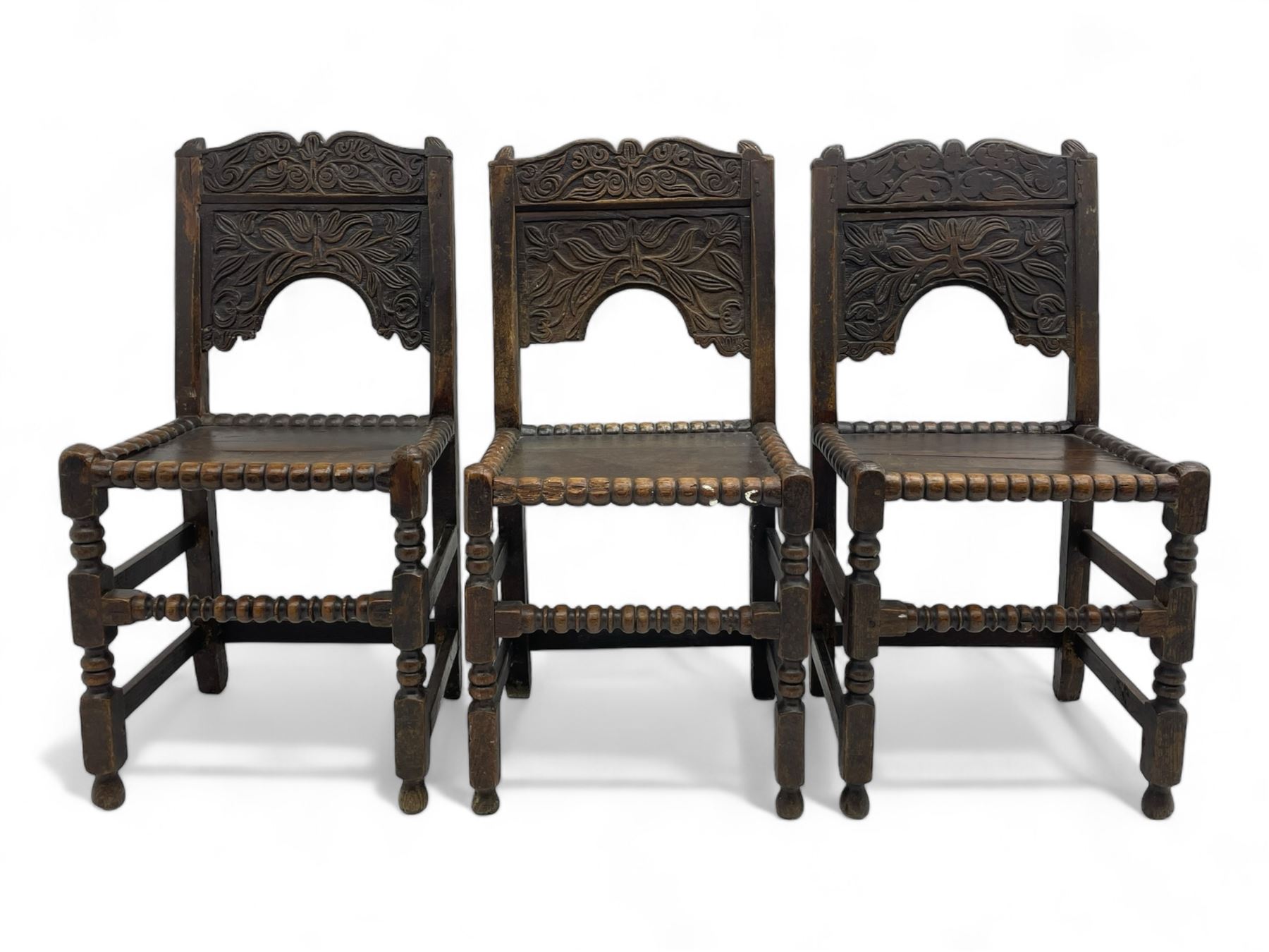 Set of three 18th century carved oak back stools, the cresting rail carved with scrolling foliate motifs over an arched panel with a stylised tree carving with extending leafage, the panelled seat held within a bobbin-turned frame, raised on turned supports united by a ring-turned front stretcher