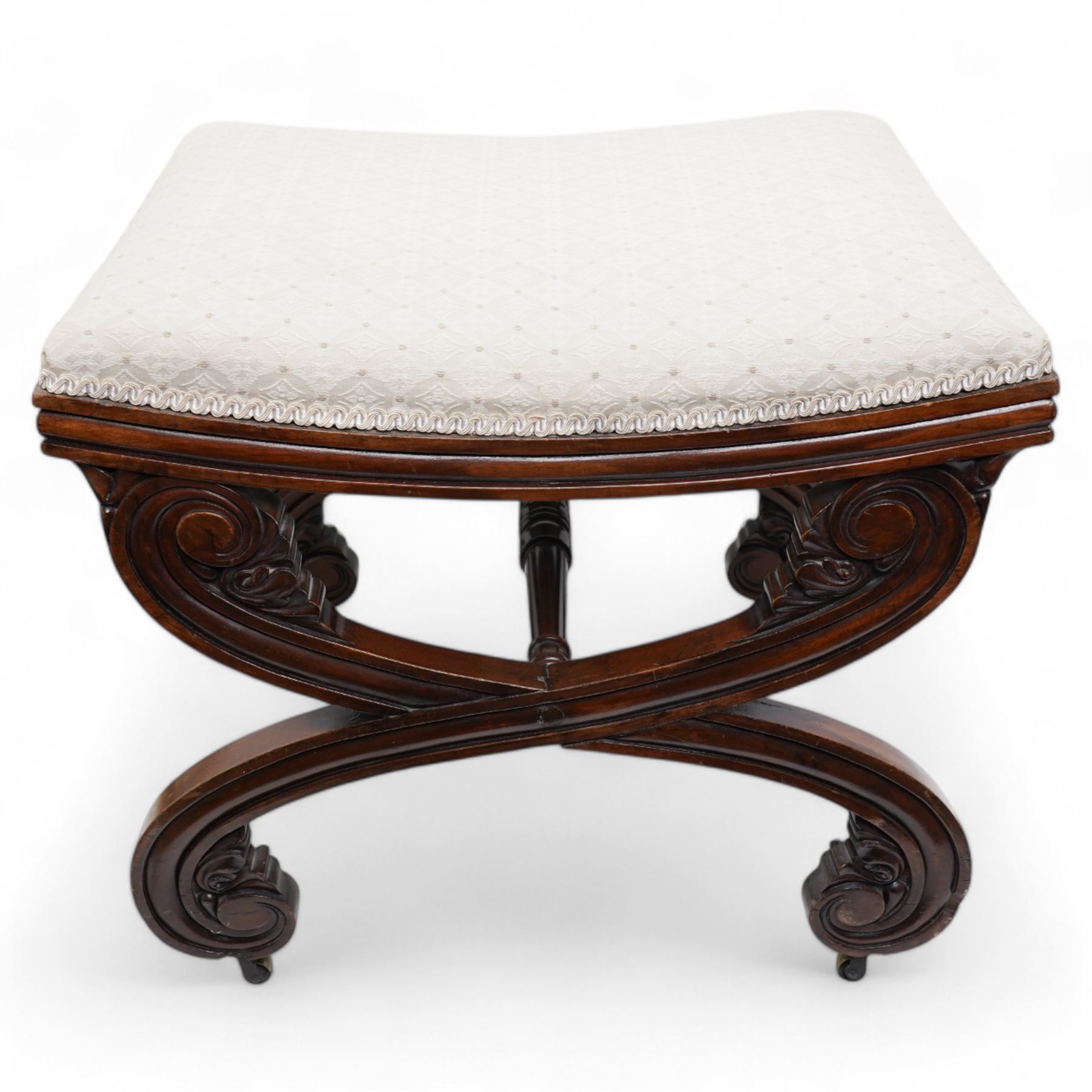 Early 19th century mahogany stool, the dished seat upholstered in cream fabric with repeating pattern, moulded seat rails, on curved X-frame supports carved with scrolls and foliage, united by turned stretcher, on brass castors