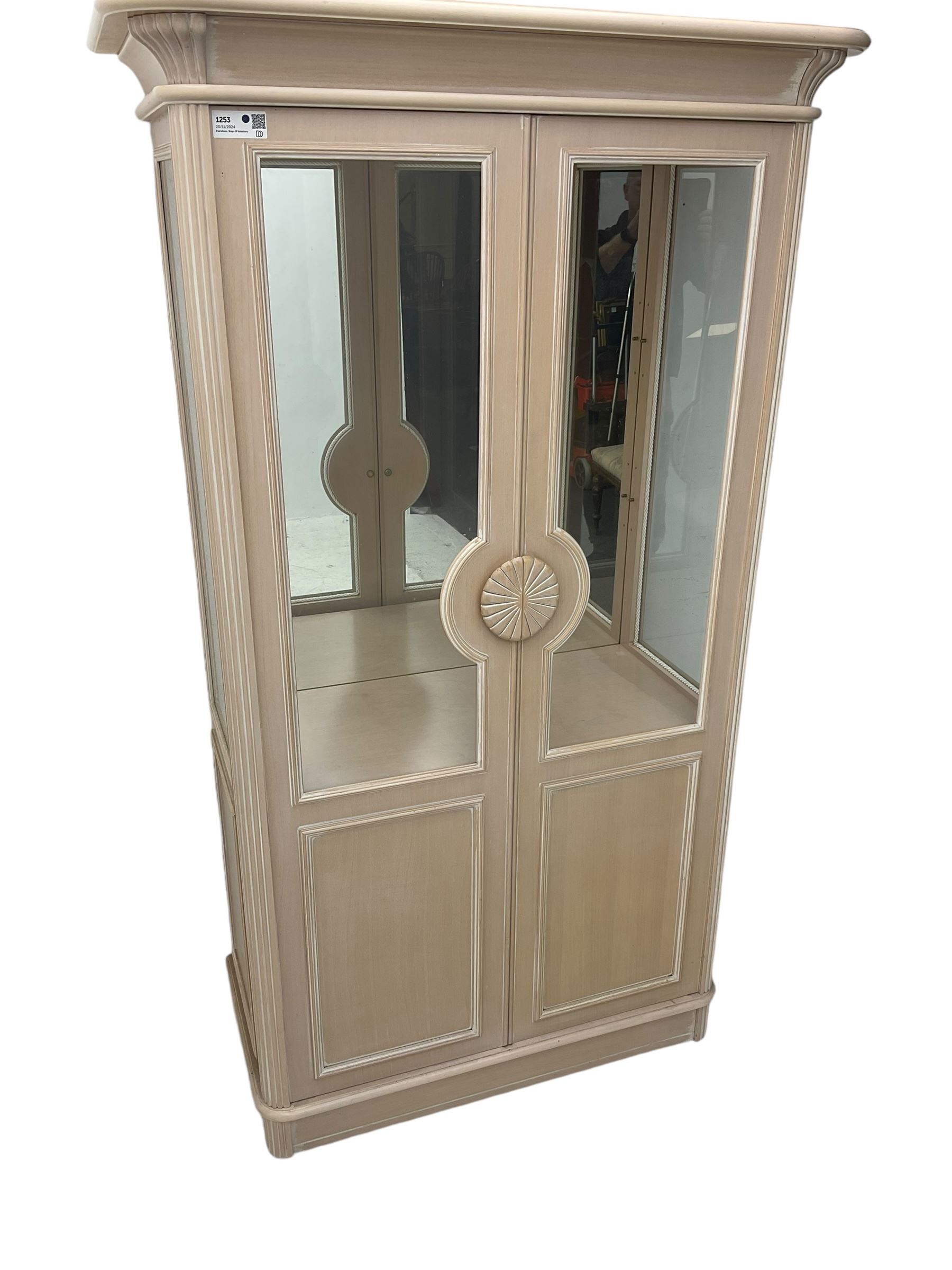 Contemporary display cabinet, moulded cornice above glazed panelled doors with central carved sunburst handles, mirrored back interior with lighting, lower enclosed storage compartment, raised on plinth base