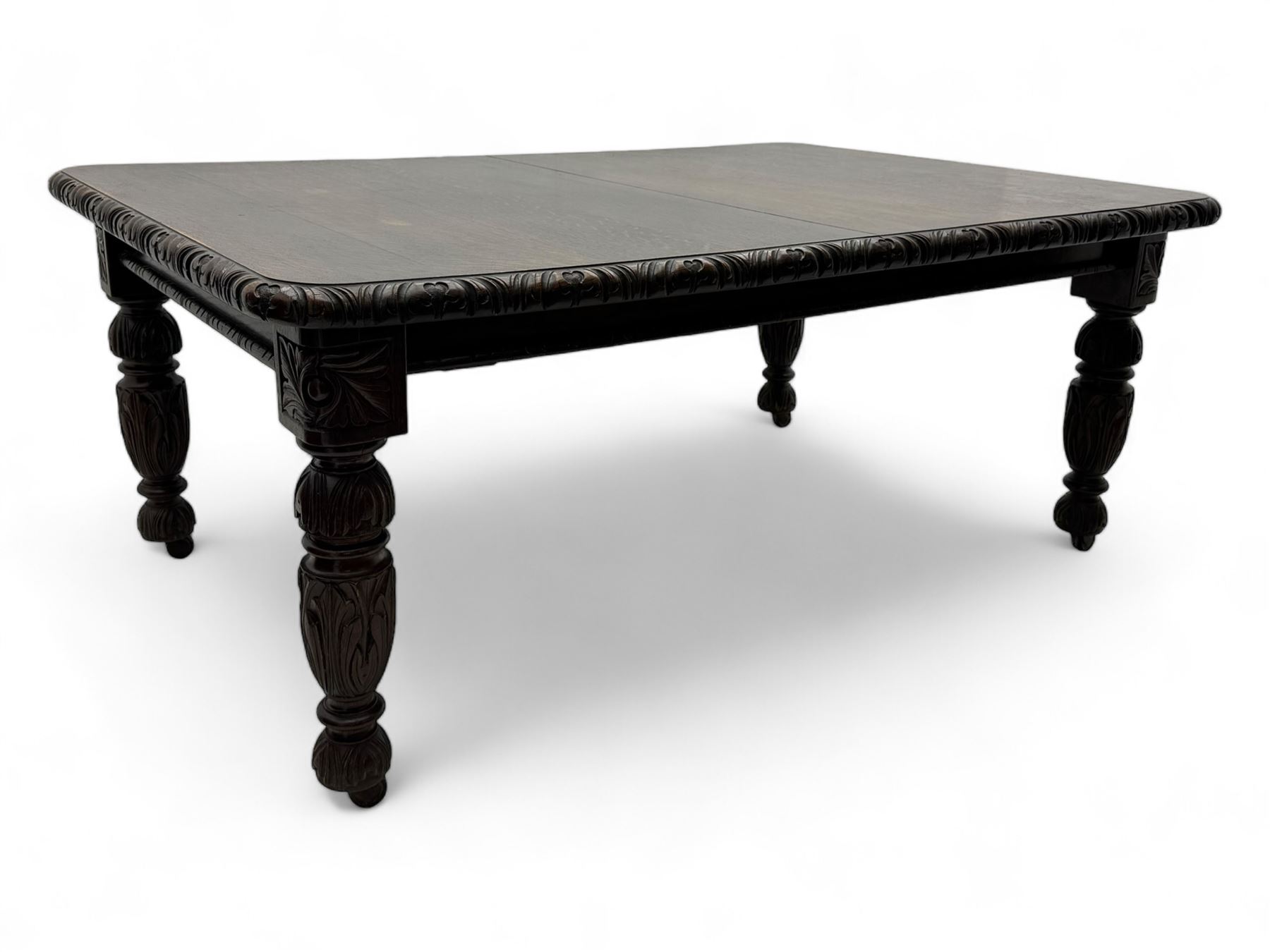 19th century heavily carved oak extending dining table, rectangular top with rounded corners and carved gadrooned edge, extending via winding mechanism with two additional leaves, raised on acanthus leaf-carved baluster supports terminating in ceramic castors