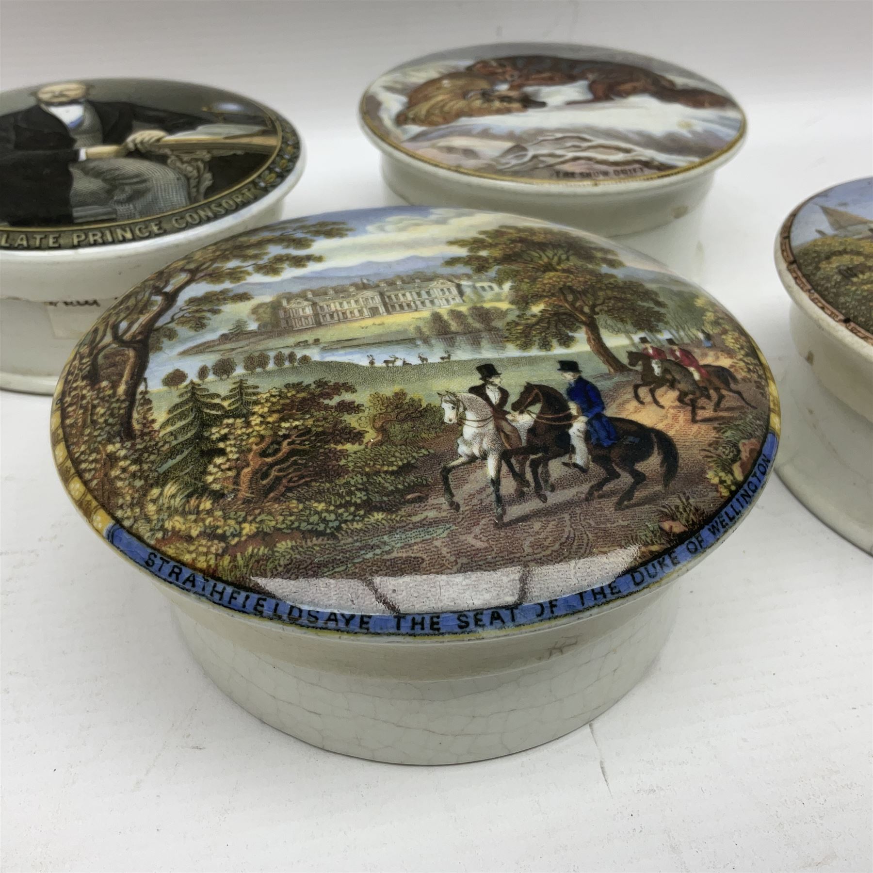 Seven 19th century Prattware pot lids with associated bases, including 'The Snow Drift', 'Strathfieldsaye The Seat of the Duke of Wellington', 'The Late Prince Consort', 'French Street Scene', Lend a Bite', 'Dr Johnson' and 'Sandringham the Seat of HRH The Prince of Wales', largest D12cm (7)