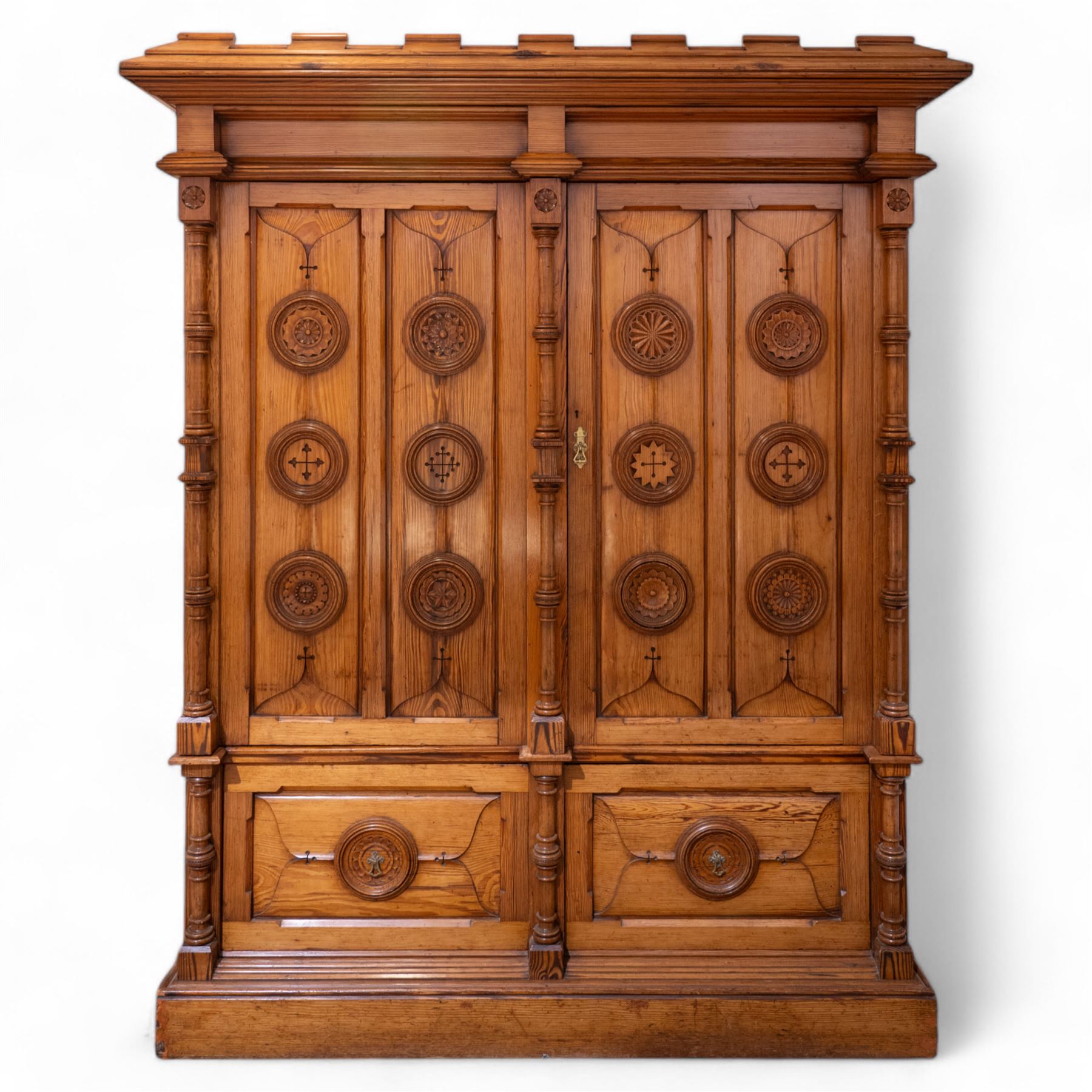 19th century pitch pine ecclesiastical vestment wardrobe or sacristy cabinet, crenelated and projecting moulded cornice over two panelled doors, the panelled doors carved with linen folds and mounted with rows of roundels depicting crucifixes and flower heads, enclosed by turned pilasters, two drawers to the base, on moulded plinth base 