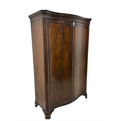 Early 20th century mahogany serpentine double wardrobe, moulded cornice over two figured doors, the interior fitted with hanging rail and shelf, canted and fluted uprights, on bracket feet 