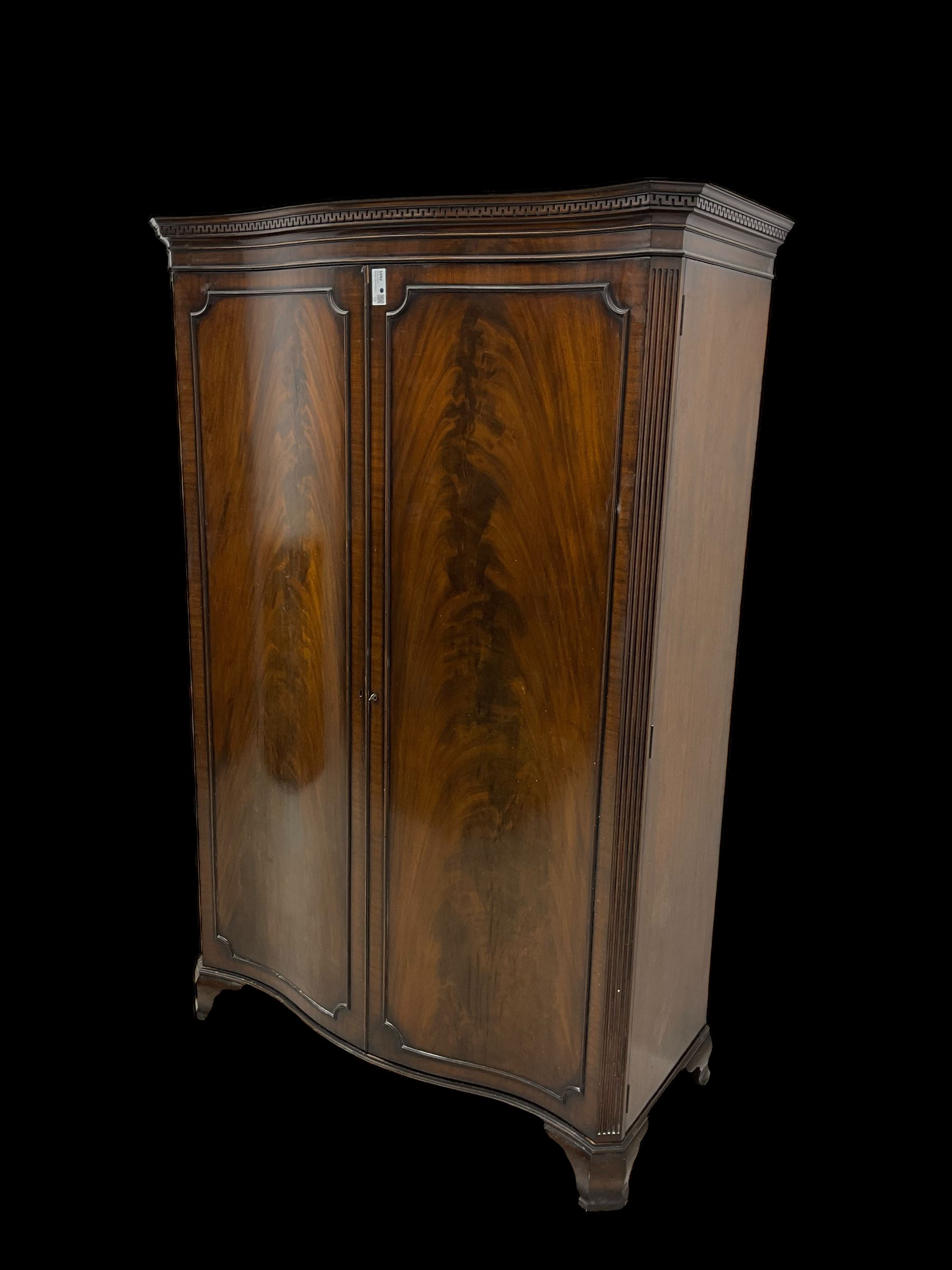 Early 20th century mahogany serpentine double wardrobe, moulded cornice over two figured doors, the interior fitted with hanging rail and shelf, canted and fluted uprights, on bracket feet 