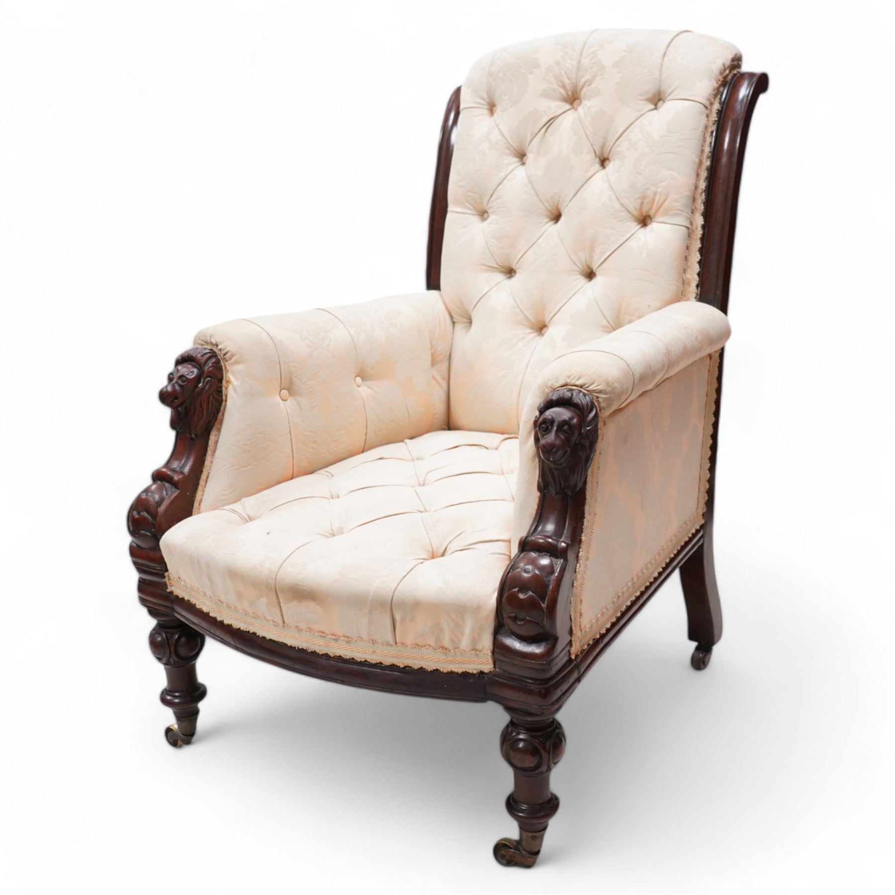 Victorian mahogany framed armchair, moulded and rolled uprights, upholstered in buttoned cream foliate pattern fabric, carved lion mask arm terminals, on turned and globular carved front feet, brass castors 