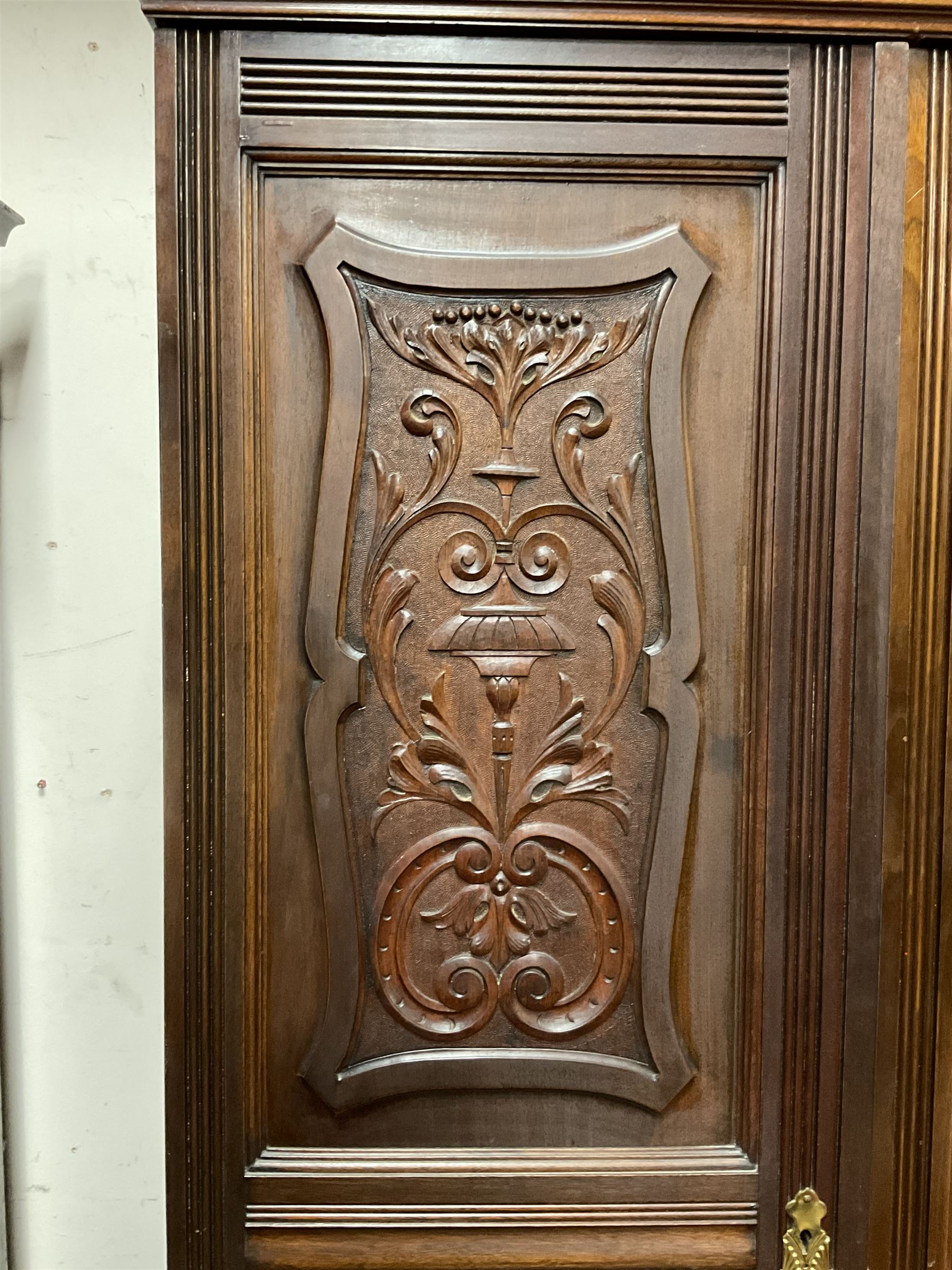 Late Victorian walnut triple wardrobe, the pierced broken swan neck pediment carved with foliate C-scrolls, central bevelled mirror door flanked by two panelled doors, the panels carved with urns, C-scrolls and trailing foliage, the interior fitted with hanging rails, long and short drawer, on moulded plinth base