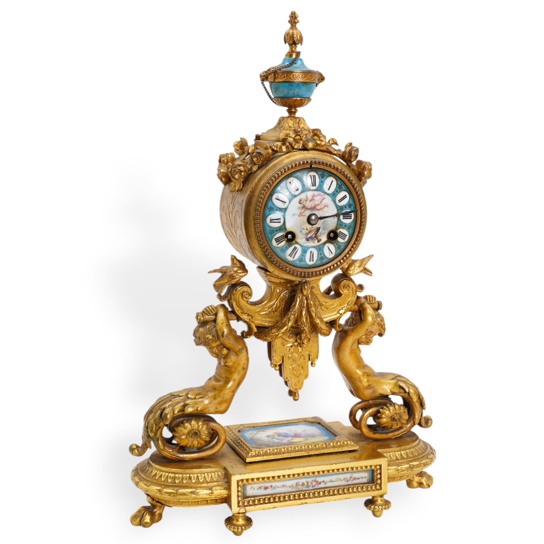 French-mid 19th century 8-day gilt and porcelain mantle clock with a break front plinth raised on paw feet, with recessed rectangular porcelain panels depicting two lovers and floral decoration, drum cased movement with a porcelain dial supported on two matching scroll supports, surmounted with a tapered porcelain urn and gilt finial, dial with cartouche Roman numerals on a blue ground and a depiction of cupid to the dial centre, twin train rack striking movement, striking the hours and half hours on a bell. With Pendulum and key.