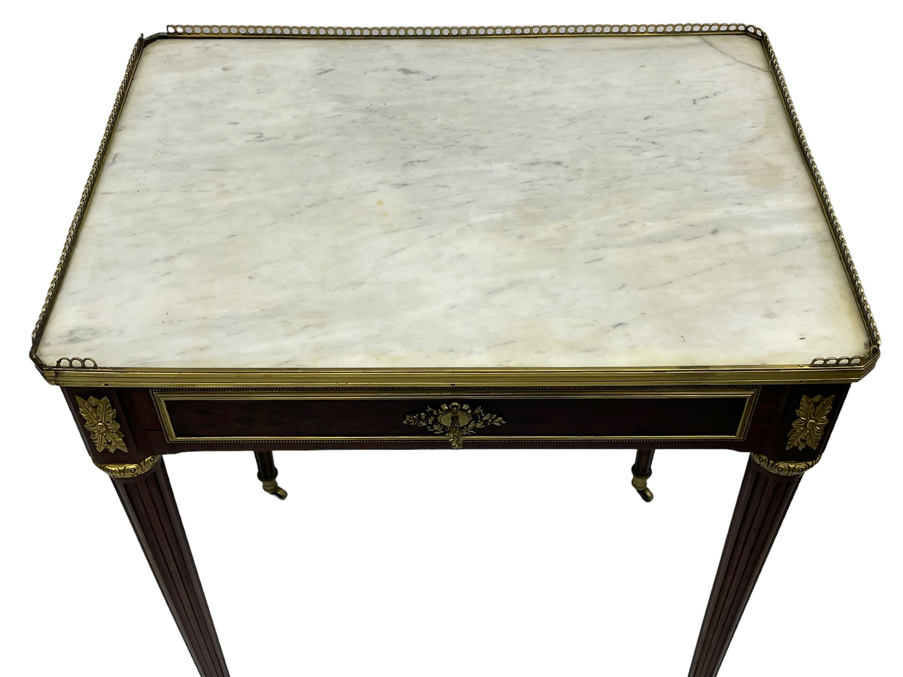 Late 19th century French figured plum pudding mahogany side table, canted rectangular white marble top with raised gilt metal gallery, fitted with a single frieze drawer, central extending floral cast ormolu escutcheon and applied moulded edging, on tapering turned and fluted supports with brass cups and castors, decorated with cast foliate mounts and foliate cast collars 