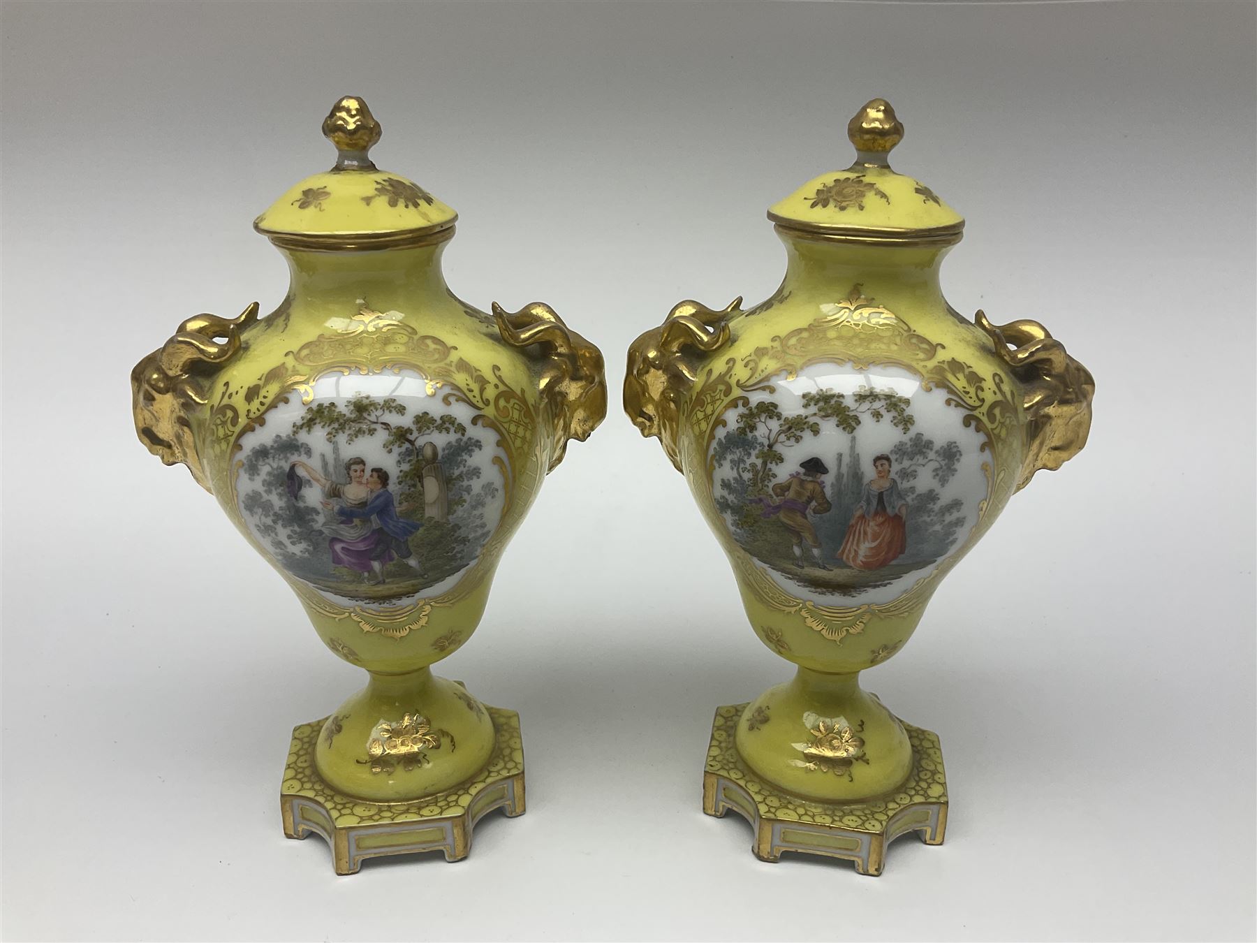 Pair of early 20th century Dresden vases with cover, of baluster form, decorated with figural panels within ornate gilt borders upon a yellow ground, with twin gilt goat mask handles, H16.5cm