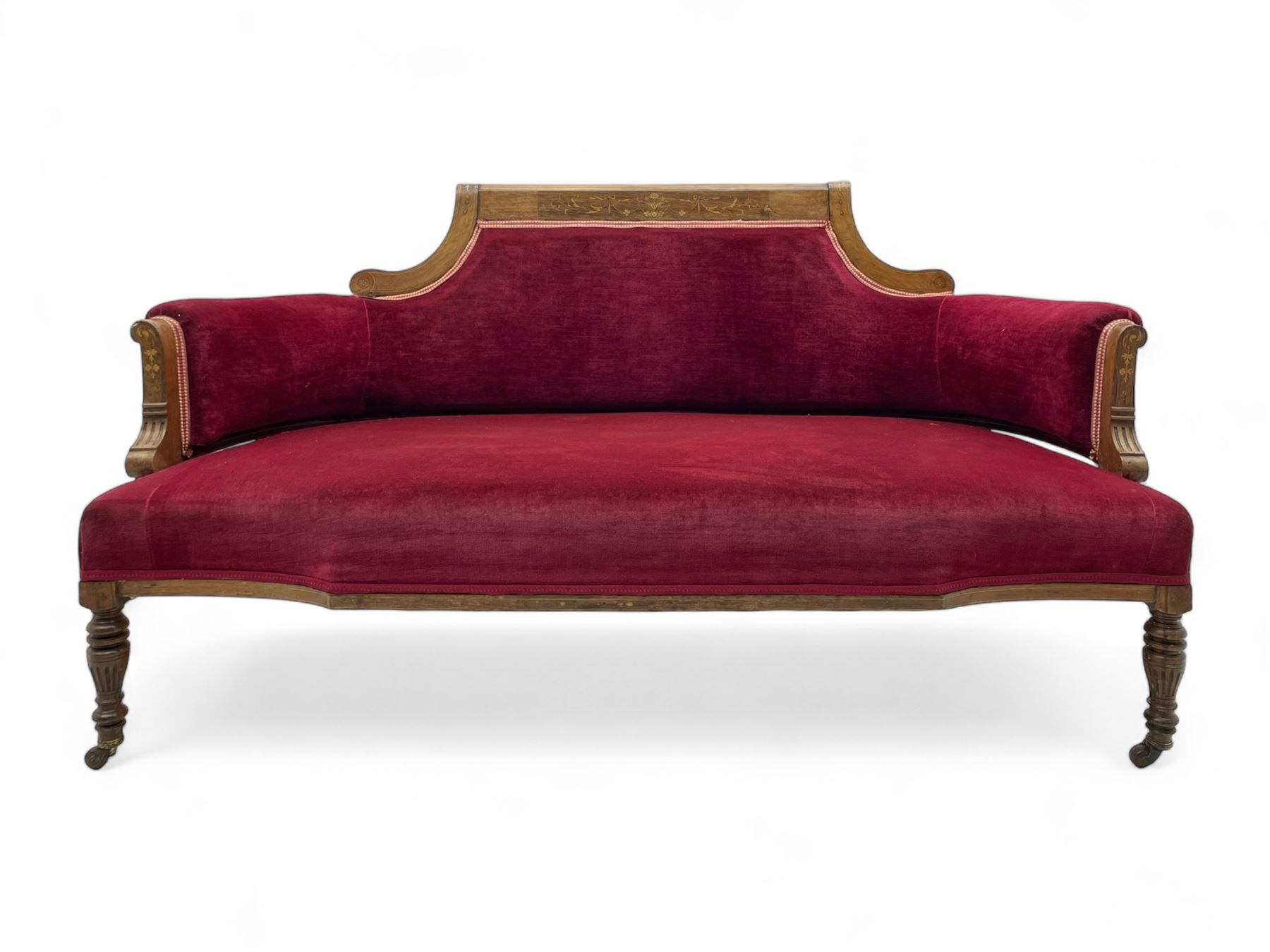 Late Victorian inlaid walnut two seat settee, the shaped cresting rail decorated with foliate inlays and boxwood stringing, upholstered in crimson velvet with sprung seat, raised on turned and fluted supports with ceramic castors