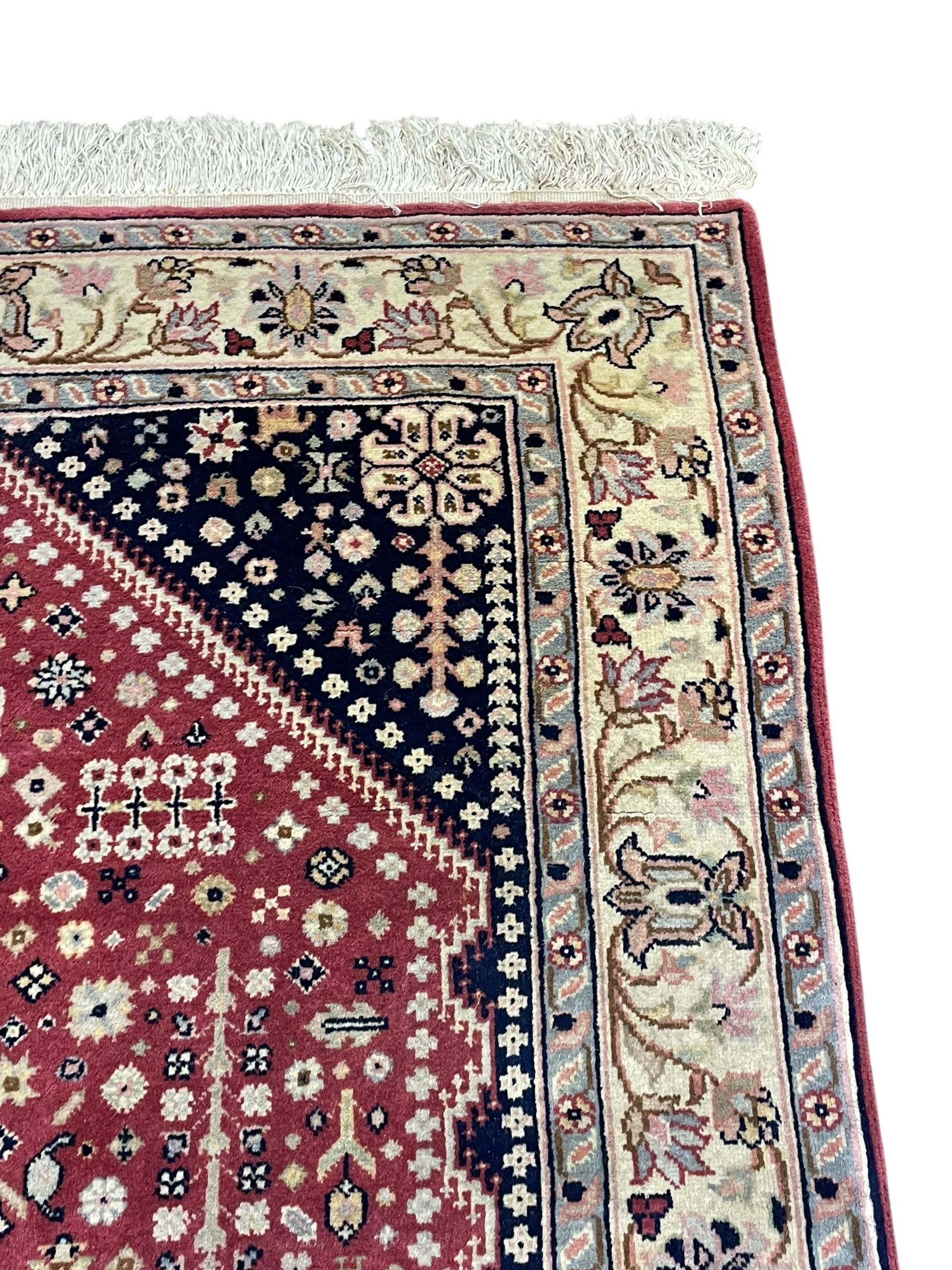 Persian design crimson ground rug, the field decorated with tree of life and floral motifs, enclosed by indigo spandrels with stylised flower head decoration, repeating scrolling border 