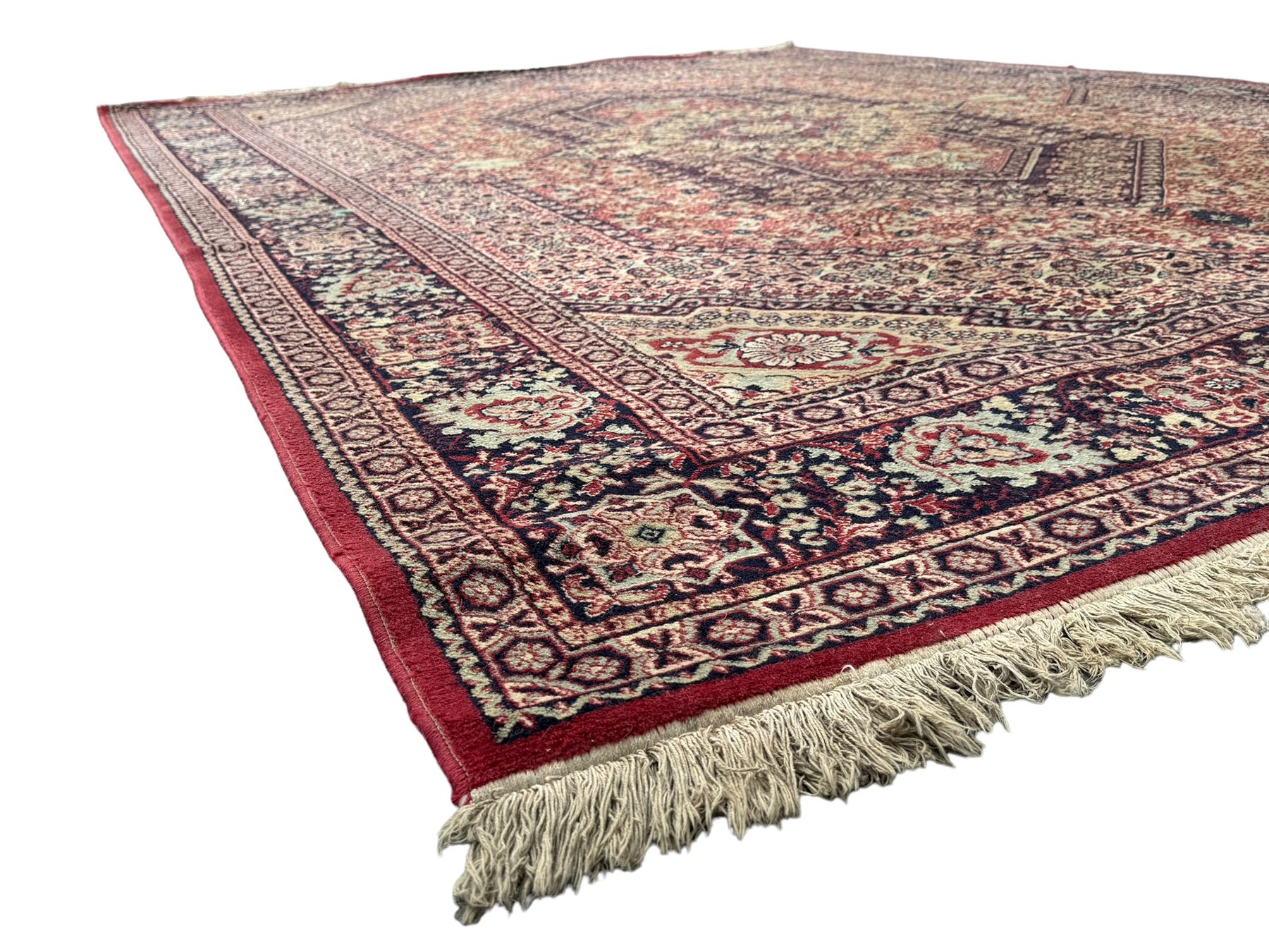 Persian Heriz design red ground carpet, the field decorated with a central star-shaped medallion surrounded by geometric floral motifs and angular vine patterns, the spandrels highlighted with palmette designs, the main border featuring a series of stylised rosettes and leaves against a dark blue ground, enclosed by multiple guard stripes with alternating floral and geometric motifs