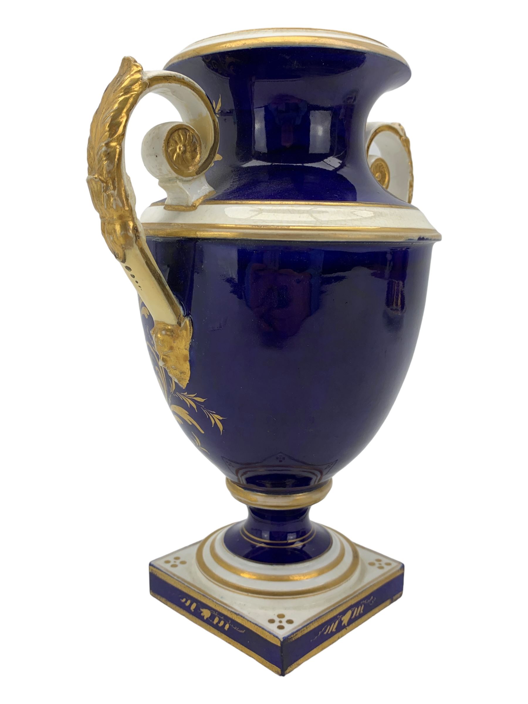 Early 19th century Derby porcelain urn, the reserve painted with a pastoral landscape and castle in the distance, with twin scroll and mask handles, upon a square pedestal base, H23cm, together with a Derby shell-shaped dish, centrally hand painted with a landscape scene titled 'In North Wales', within a cobalt blue and gilt border, L25cm (2)