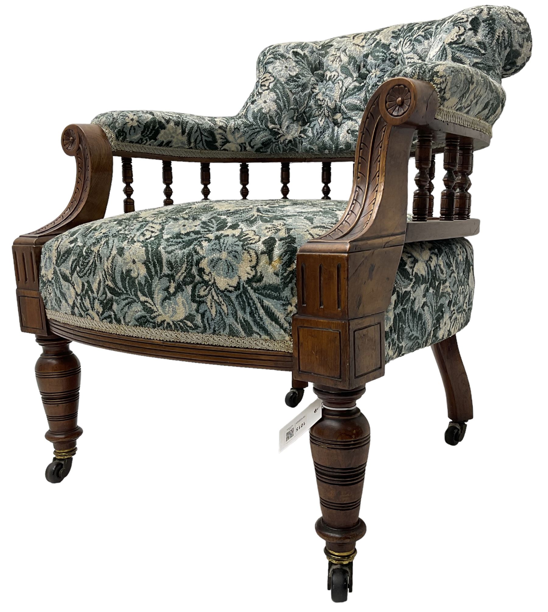 Victorian walnut framed tub-shaped armchair, the shaped rolled back supported by a spindle turned balustrade, upholstered in buttoned floral fabric, scrolled arms supports carved with acanthus leaves and flower heads, on collar turned feet with brass and ceramic castors