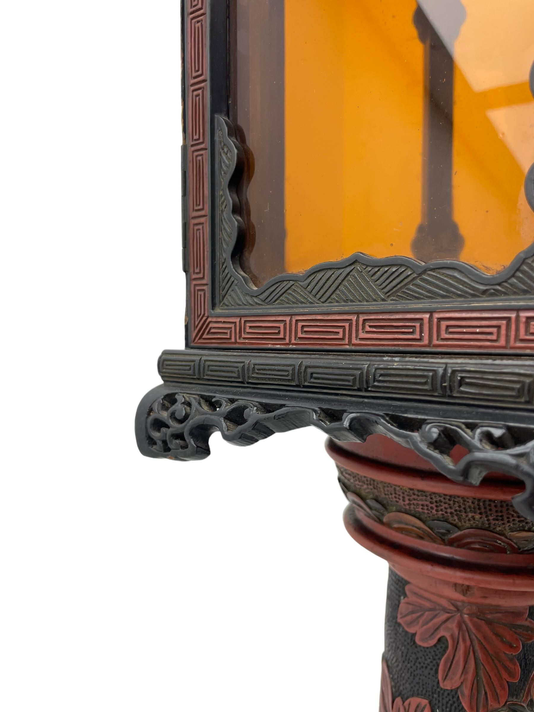 20th century Chinese black and red lacquered hardwood lantern stand, hexagonal shaped canopy top carved with flower heads and scrolls, the lantern fitted with amber tinted glass panes, pierced and carved upper panels, enclosed by two hinged doors, turned stem carved in relief with trailing vine leaves with fruits, stepped hexagonal base with carved decoration  