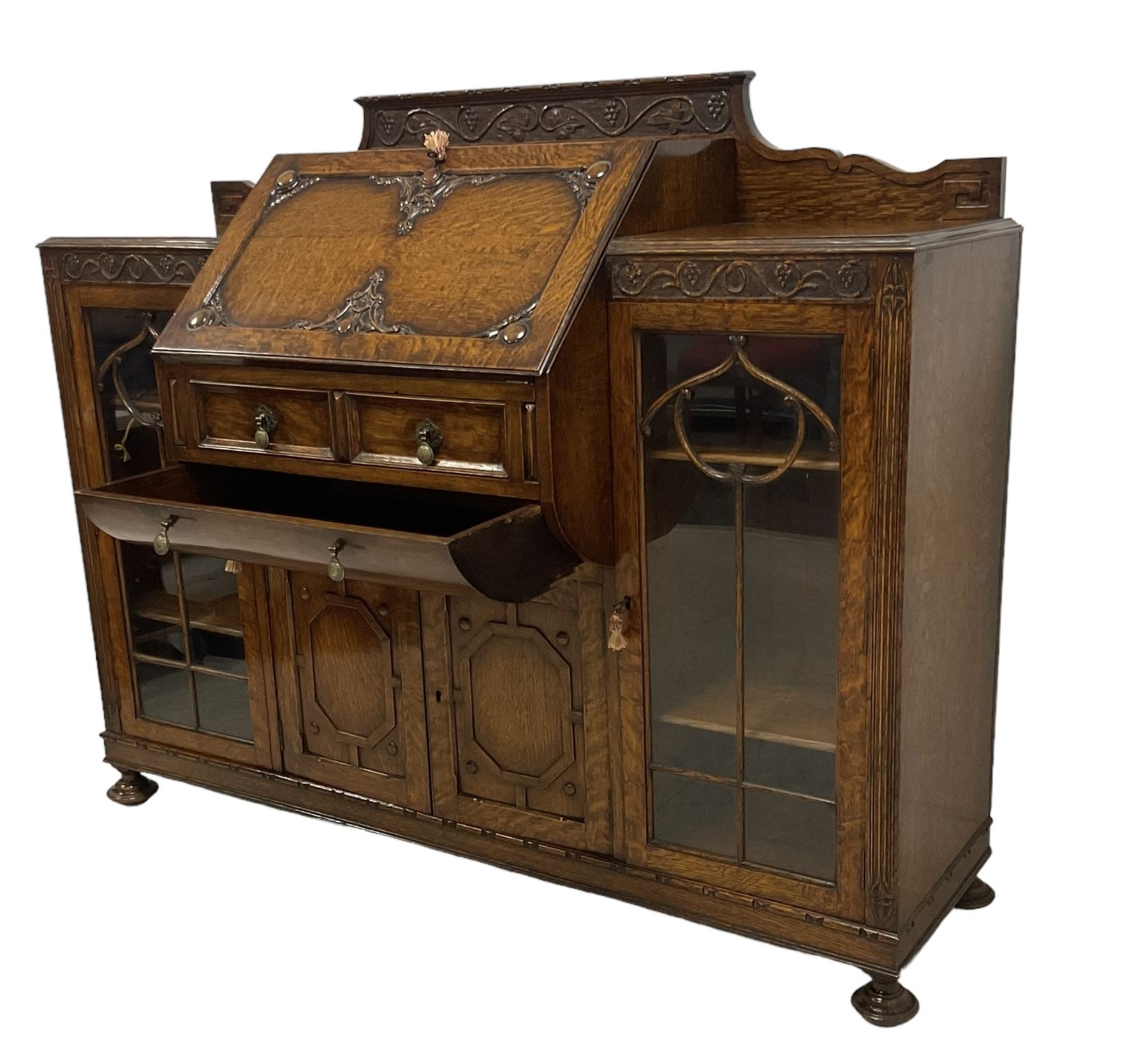 Early 20th century heavily carved oak bookcase bureau, raised back with grape-vine carved frieze, over fall-front enclosing fitted interior, above two short drawers and curved frieze drawer, flanked by astragal glazed and panelled cupboards, on turned feet