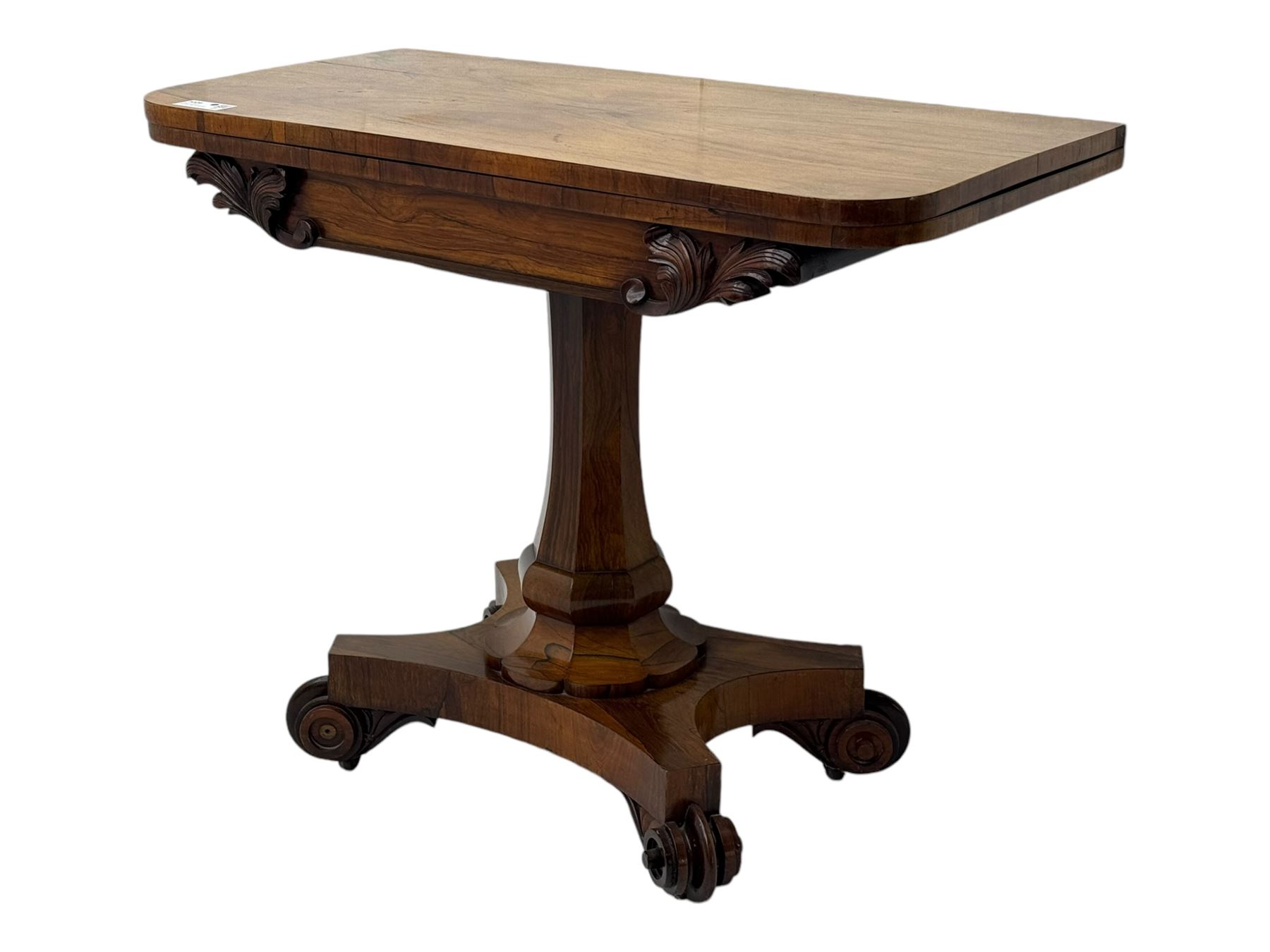 Early Victorian rosewood card table, rectangular foldover swivel top with rounded corners, red baize-lined surface, frieze adorned with carved foliate motifs, supported by a central octagonal waisted column on shaped quatrefoil base, terminating in scrolled feet with concealed metal castors