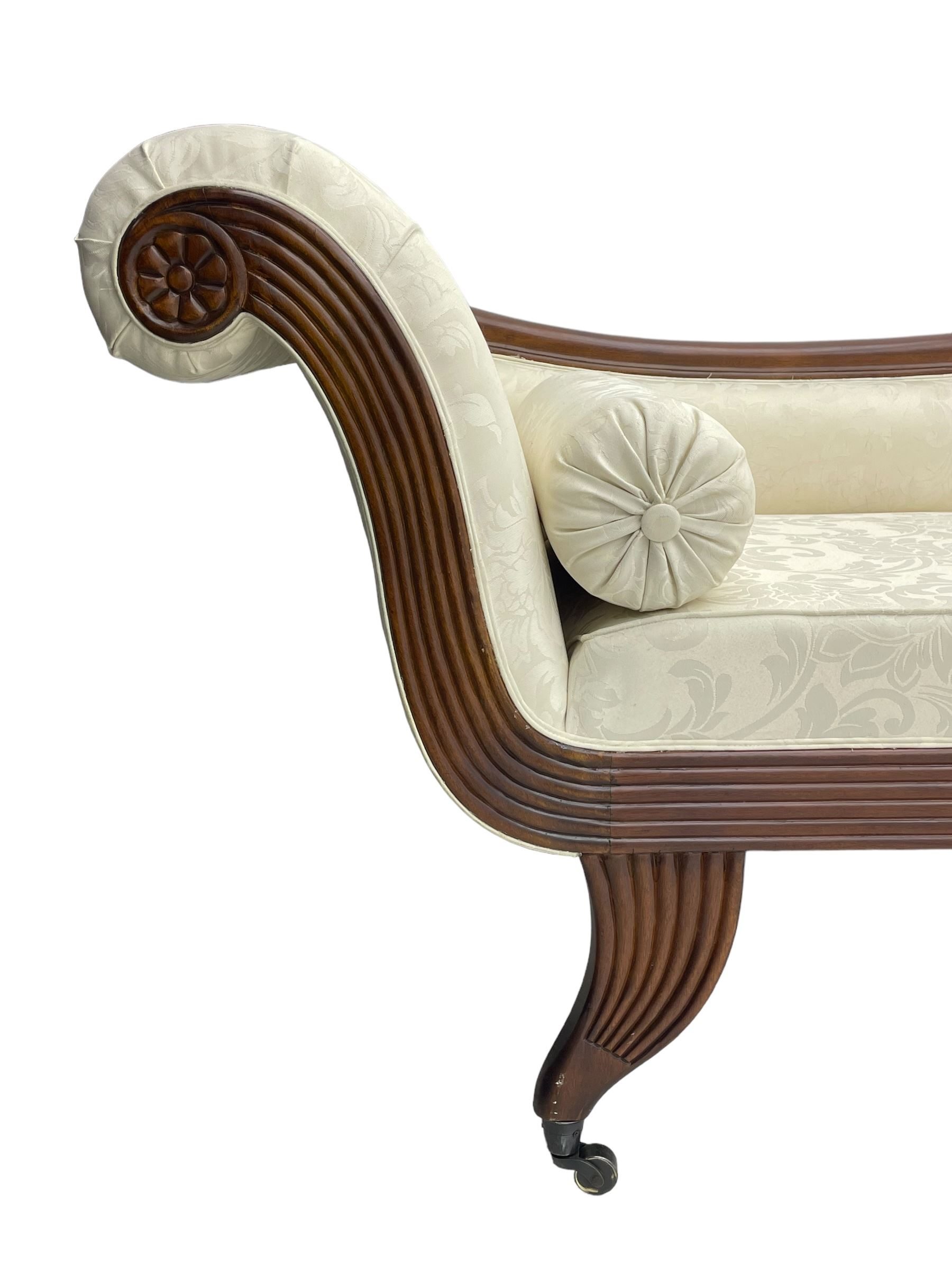 Regency design mahogany chaise longue, scrolled arms with carved rosette details, upholstered in cream damask fabric with bolster cushion, reeded frame supported by turned legs on brass castors