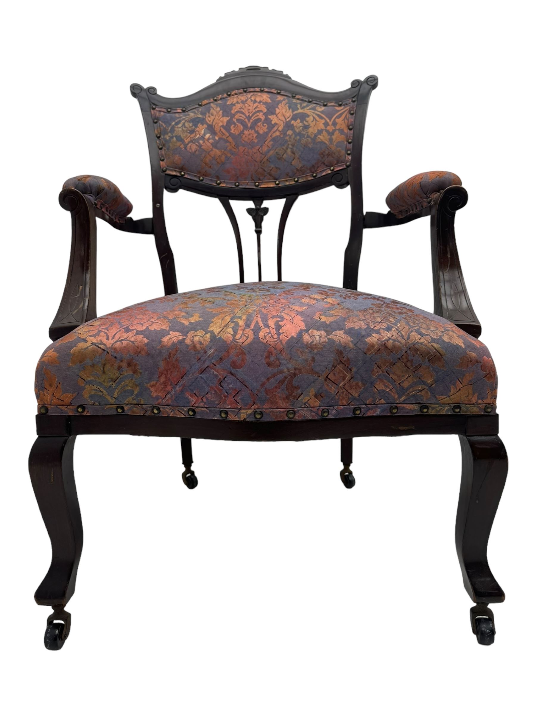 Late Victorian mahogany framed armchair, upholstered in muted purple fabric with iridescent floral pattern and nailhead trim, central foliate carved splat over padded scrolled arms, on cabriole supports fitted with castors