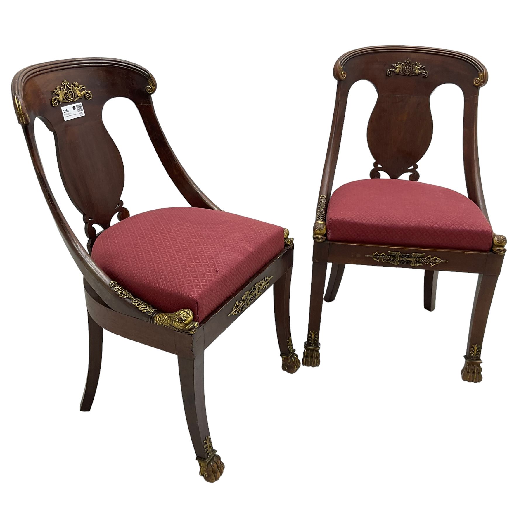 Pair of French Empire mahogany spoon0back side chairs, reeded cresting rail with scrolled ends over a shaped and pierced back splat, decorated with gilt metal mount in the form of winged classical maidens playing panpipes, drop-in seat upholstered in crimson lozenge patterned fabric flanked by gilt stylised scaled fish mounts, the apron with neoclassical gilt metal mounts, raised on sabre supports with paw feet