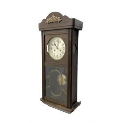 Oak cased 1930s 8-day German wall clock - with a crested top, glazed door and visible pendulum, silvered dial with Arabic numerals and spade hands, two train movement striking the hours on a gong. With pendulum and key.