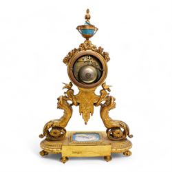 French-mid 19th century 8-day gilt and porcelain mantle clock with a break front plinth raised on paw feet, with recessed rectangular porcelain panels depicting two lovers and floral decoration, drum cased movement with a porcelain dial supported on two matching scroll supports, surmounted with a tapered porcelain urn and gilt finial, dial with cartouche Roman numerals on a blue ground and a depiction of cupid to the dial centre, twin train rack striking movement, striking the hours and half hours on a bell. With Pendulum and key.